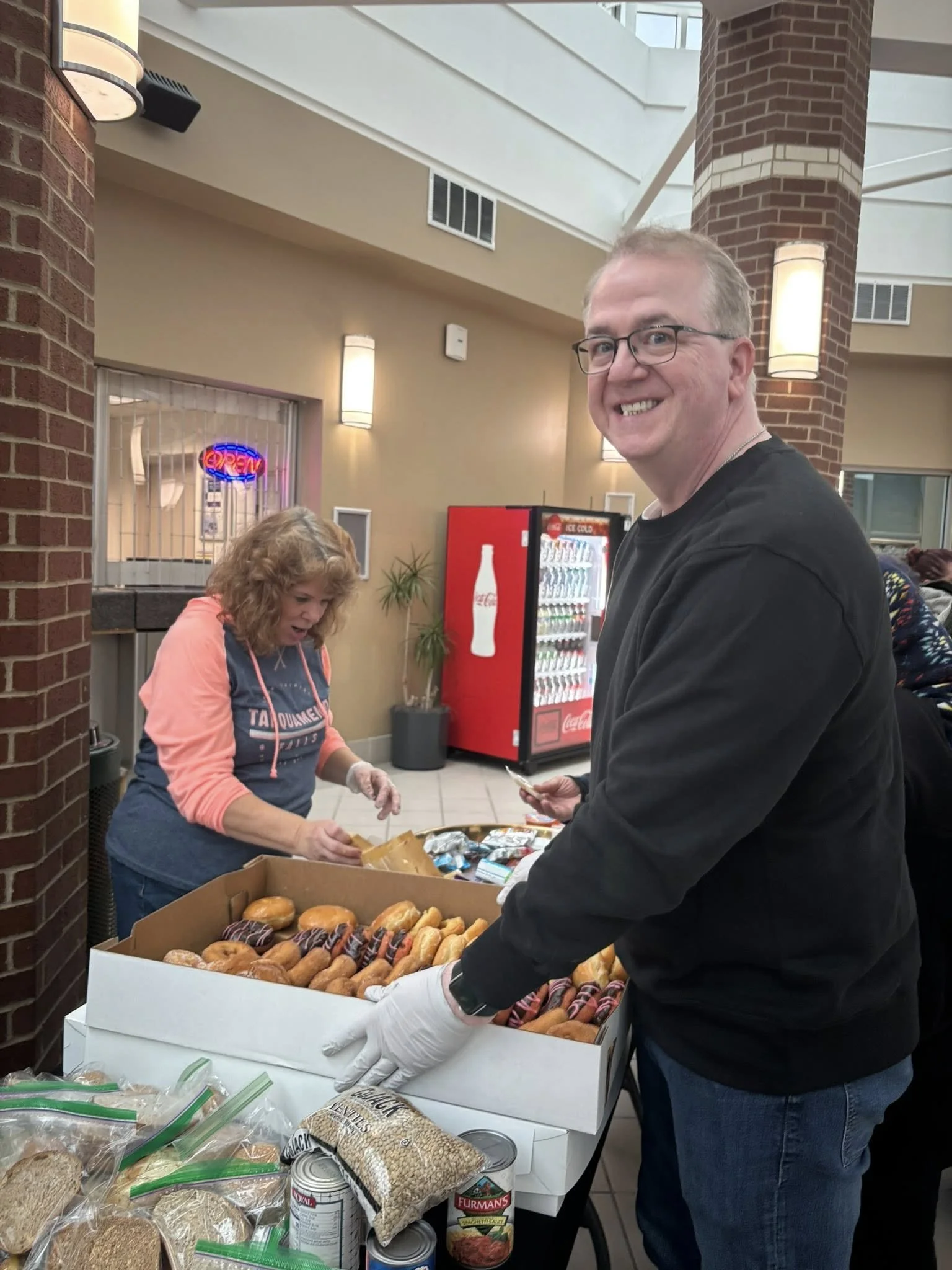 A man with glasses and a black sweatshirt smiling while serving donuts, with a woman behind him in a hoodie fetching donuts, in a room with a vending machine, potted plant, and a brick wall.