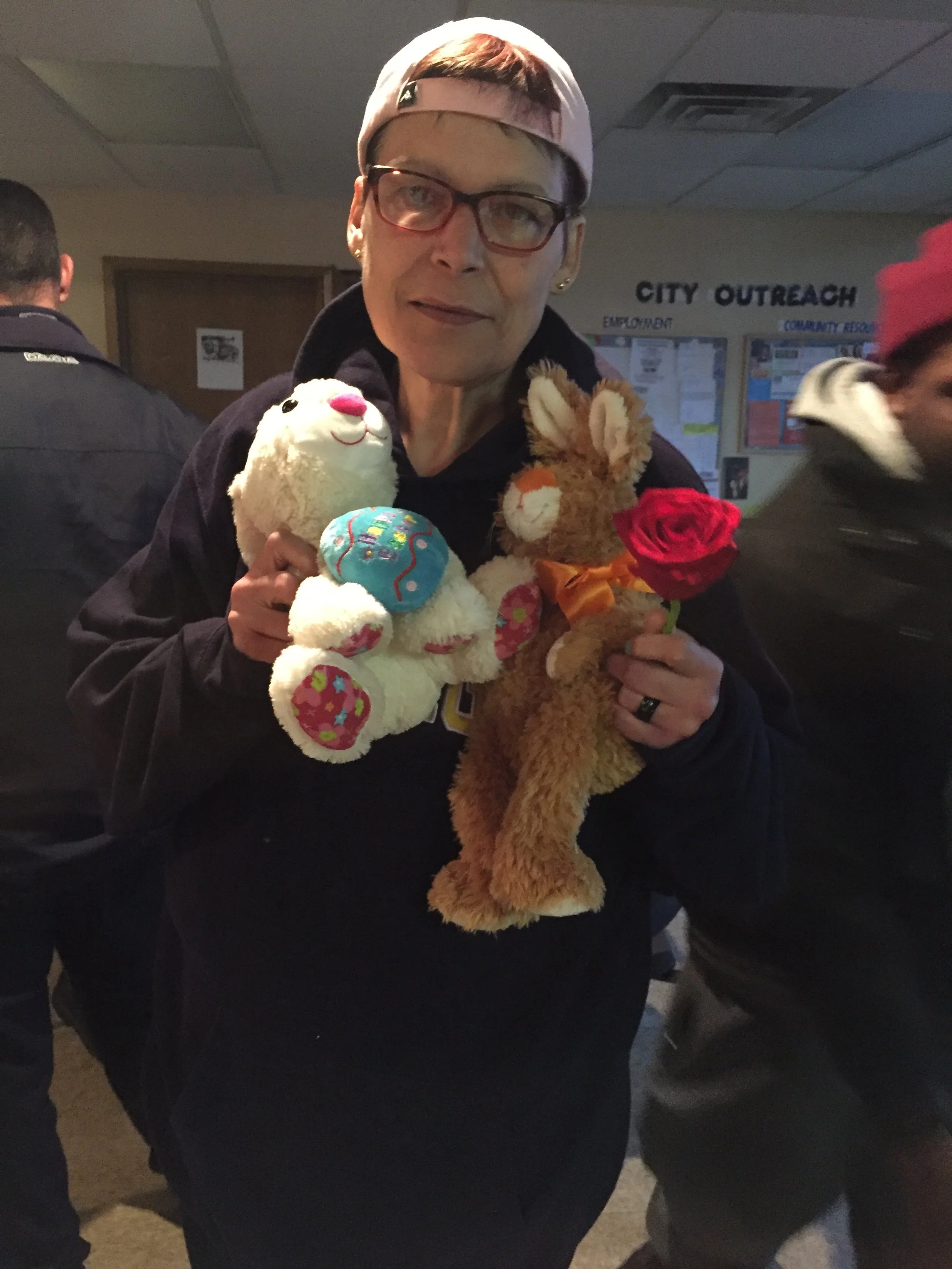 Woman wearing glasses and a baseball cap holding two plush toys, a white sheep and a brown rabbit, each with a small plush egg, and a red rose.