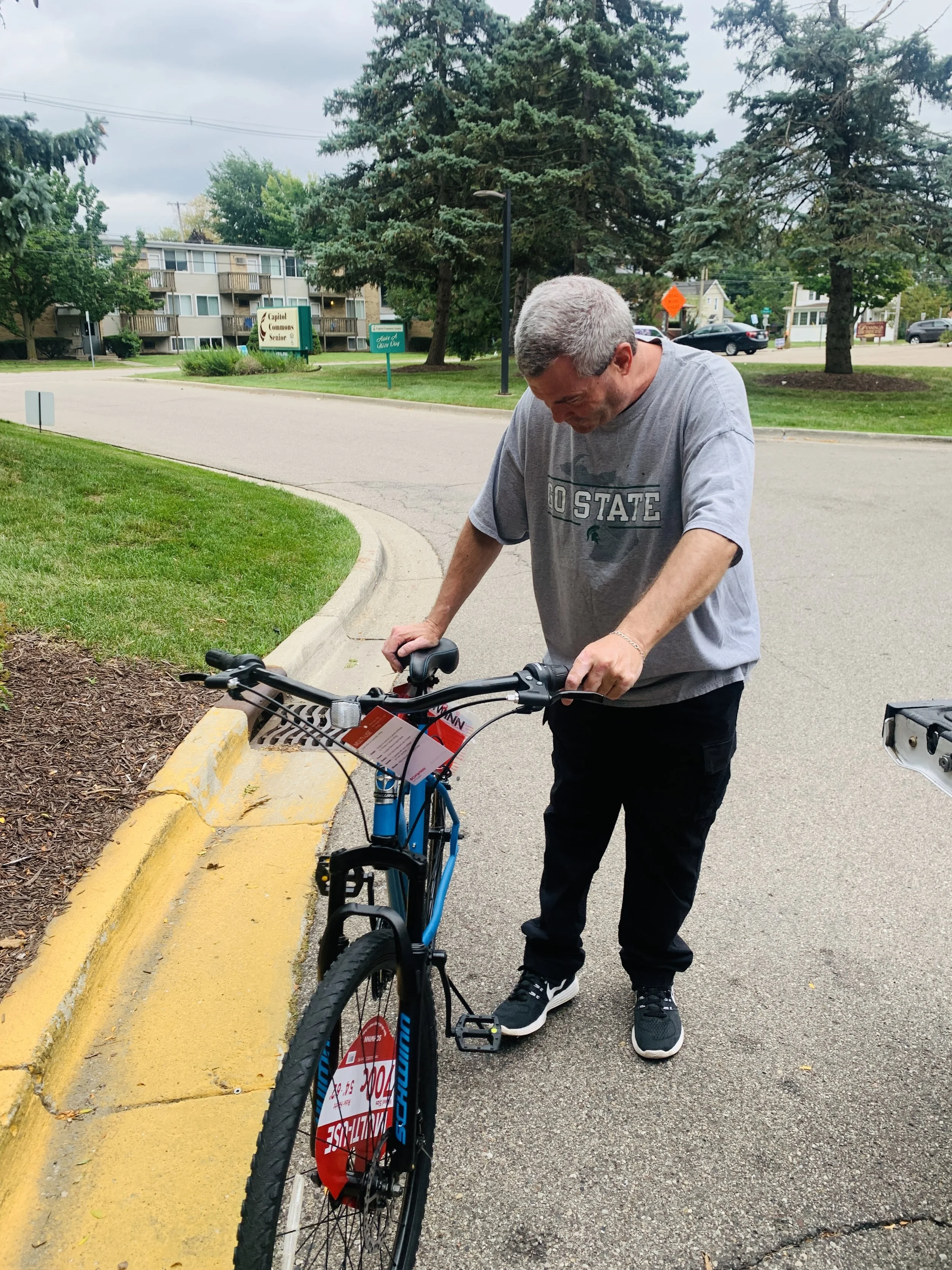 A man in a gray T-shirt and black pants is standing next to a blue bicycle on a sidewalk, leaning over to adjust or examine it.