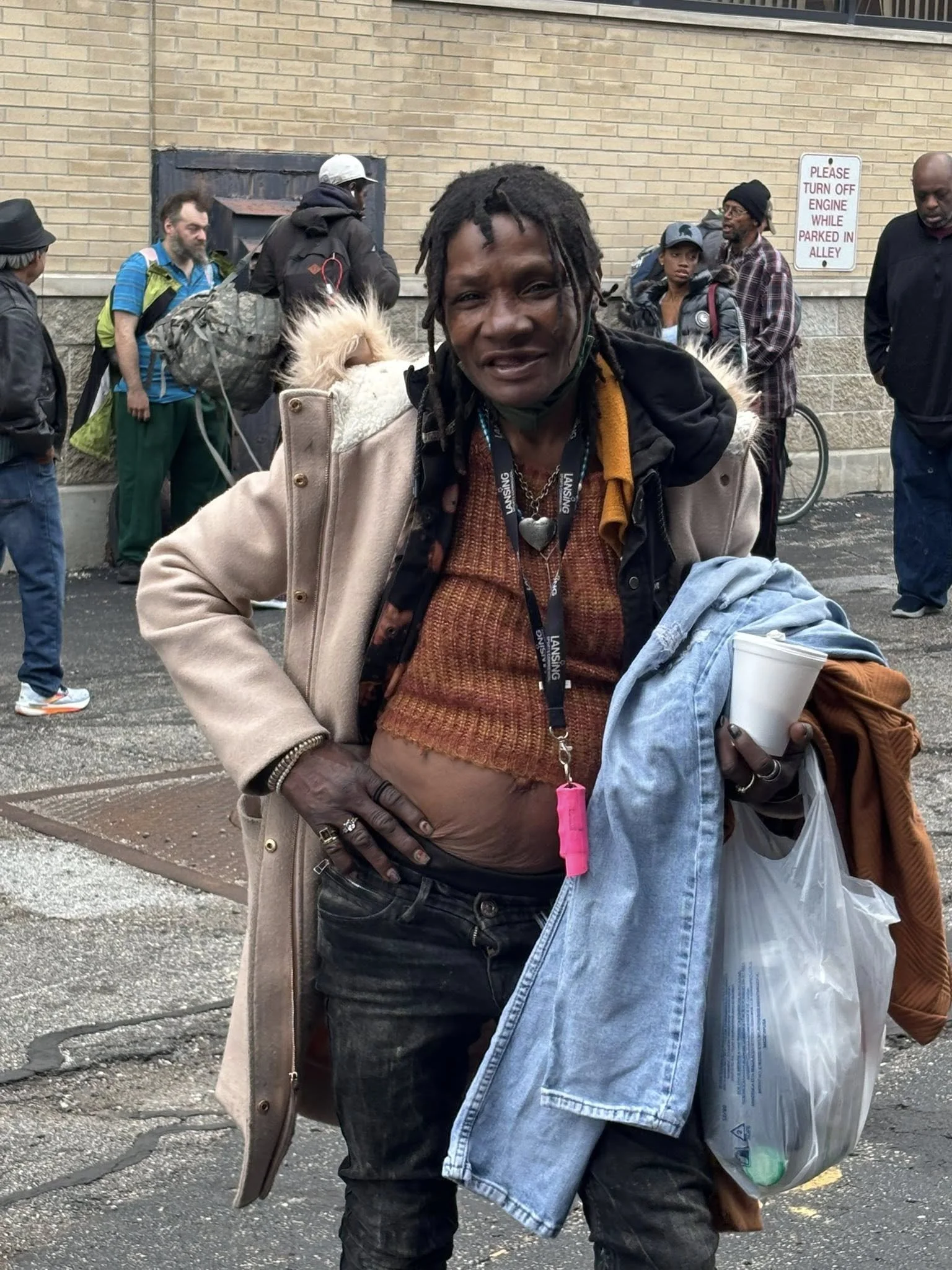 Woman with dreadlocks smiling, wearing a beige jacket, holding clothing and a cup, standing outside among people.