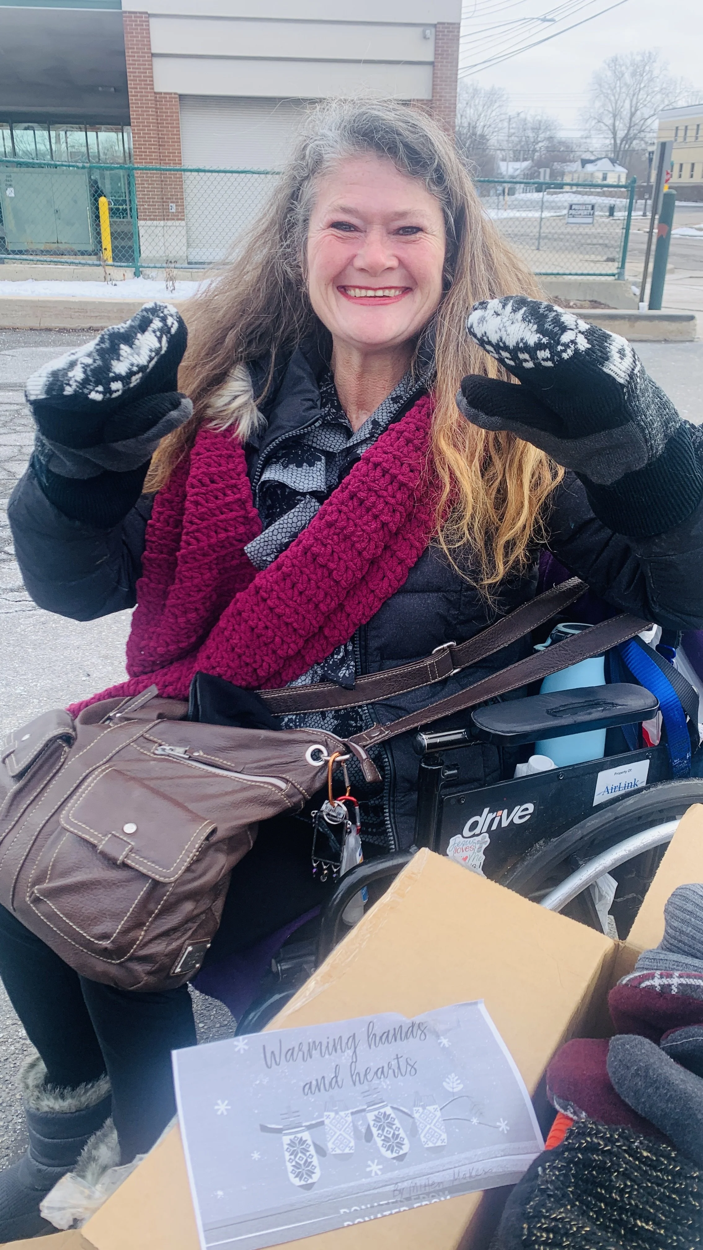 A smiling woman in winter clothing, including gloves, scarf, and jacket, standing outdoors next to a wheelchair and a donation box with a sign that says 'Warming hands and hearts. Warm.' in a parking lot with a chain-link fence and buildings in the b