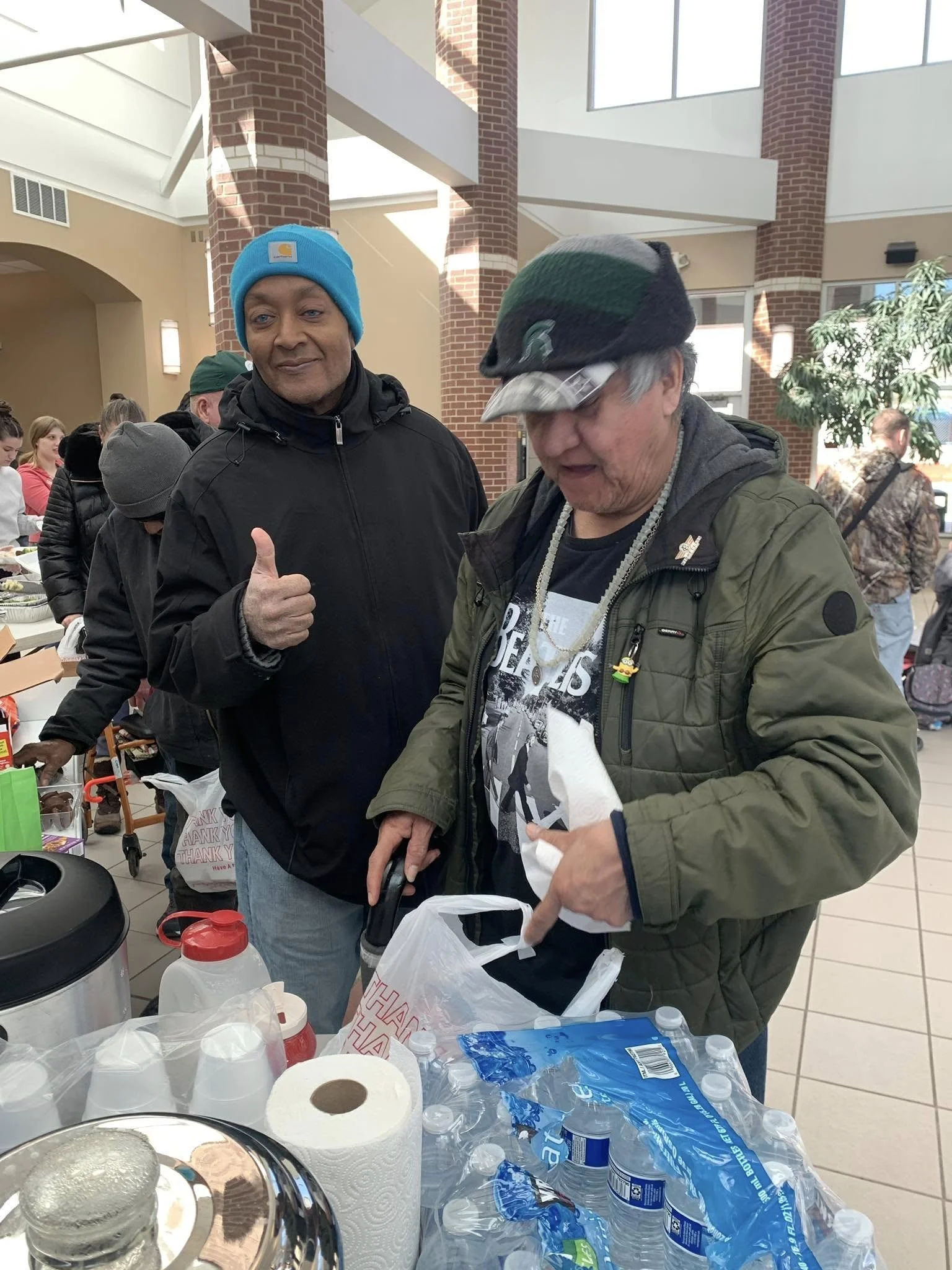 Two men standing at a table filled with food and beverages indoors. One man is giving a thumbs-up and is wearing a blue beanie and black jacket. The other man is wearing a green jacket, a cap, and glasses, looking down at a plastic bag. There are other people and tables in the background.