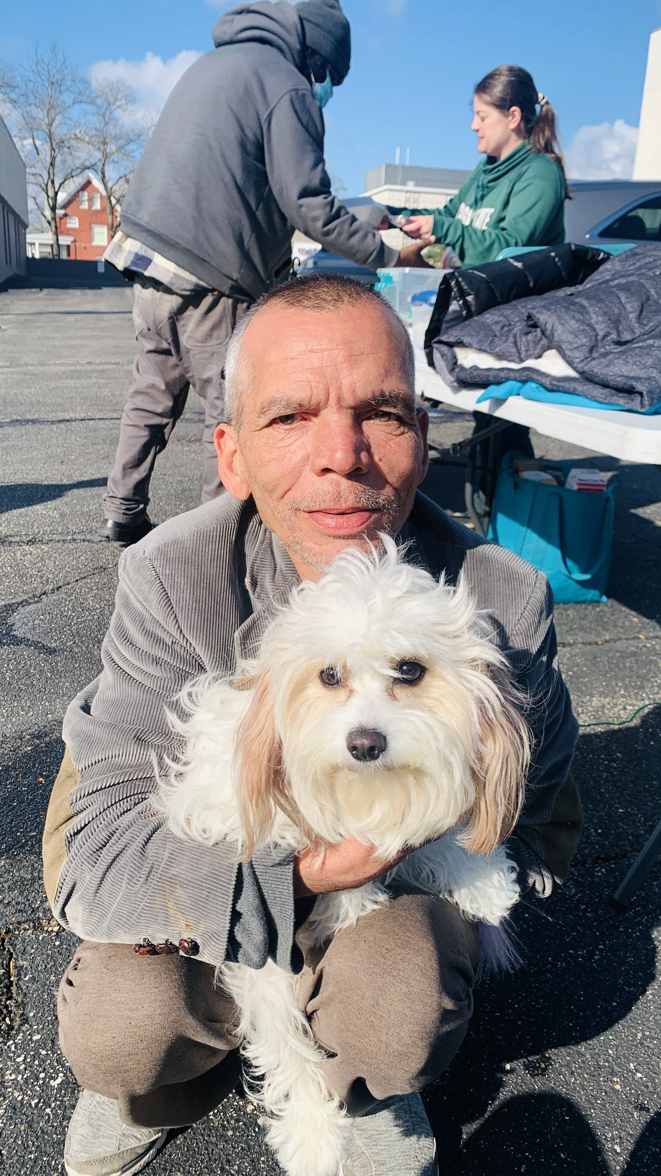 Man crouching down holding a fluffy white dog outdoors on a sunny day, with a table and two people in the background.
