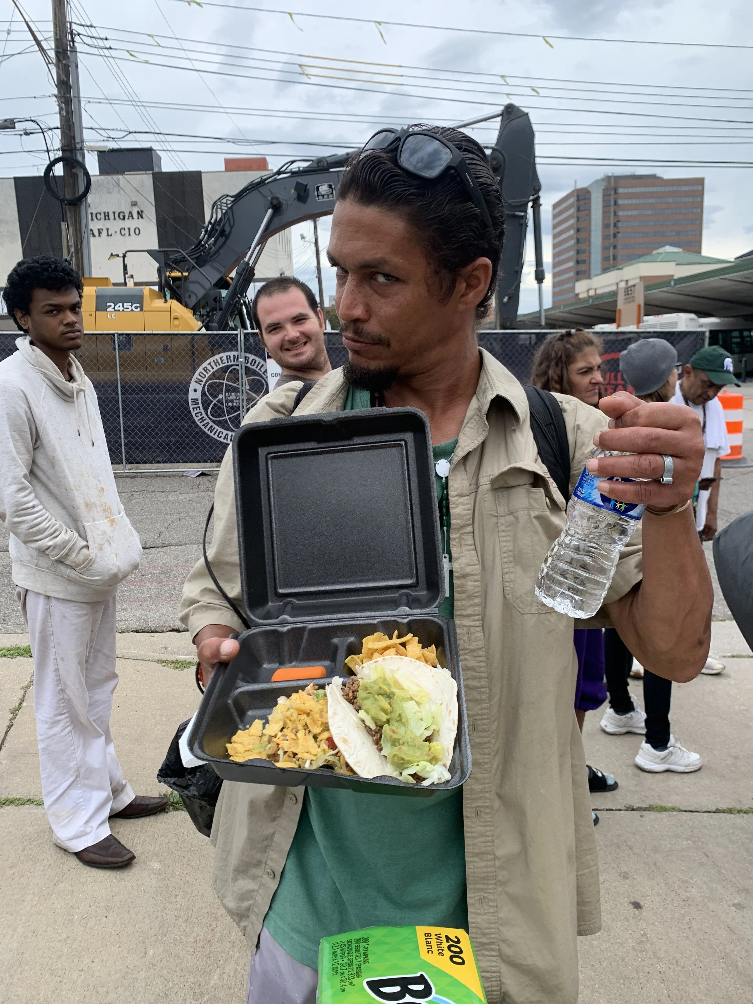 A man holding a takeout container with food including tacos and chips, and a bottle of water. He is wearing sunglasses on his head and has a serious expression. There are multiple people and construction equipment in the background, outdoors on a clo