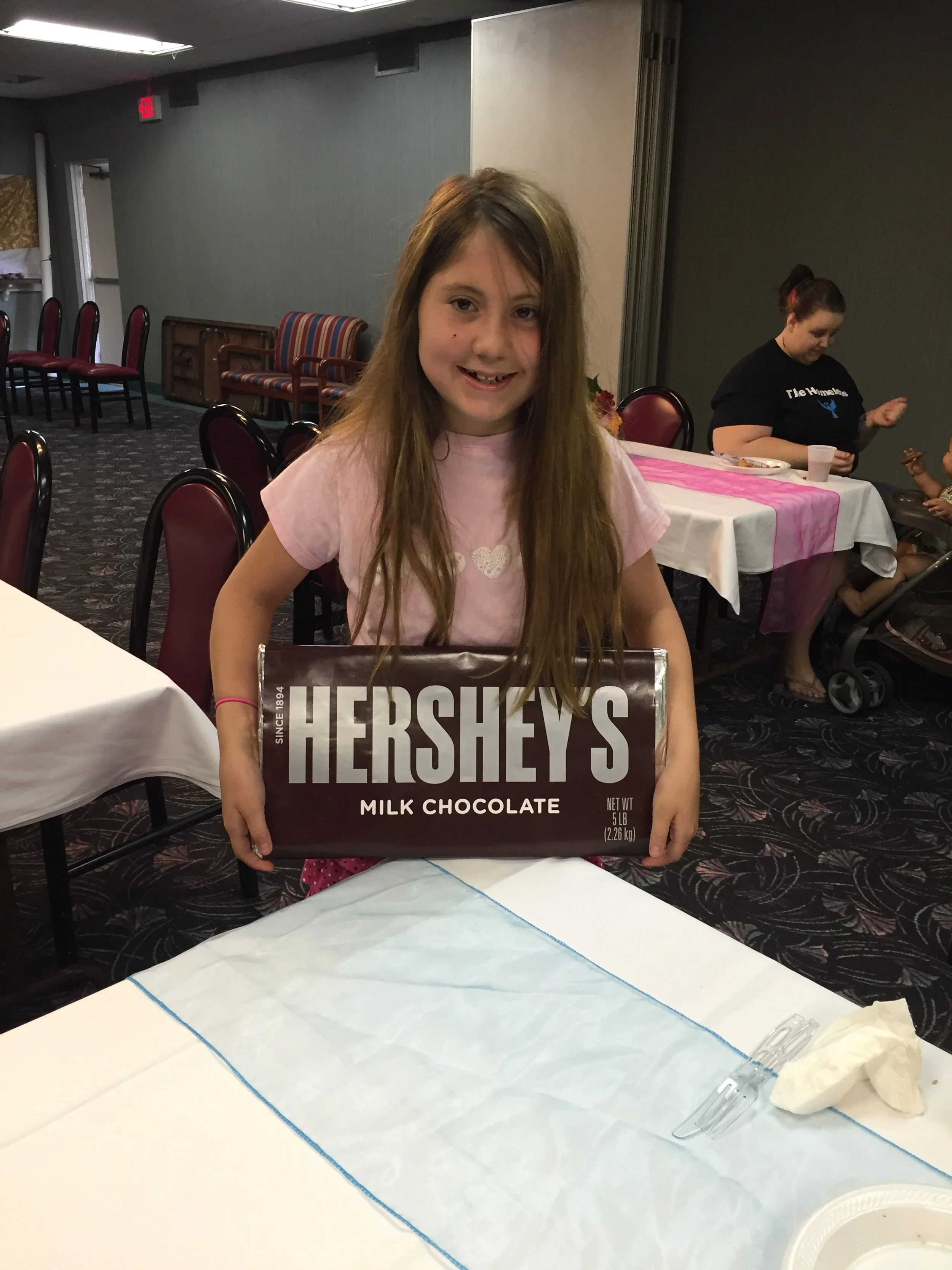 A young girl with long brown hair wearing a pink shirt, smiling and holding a large Hershey's milk chocolate bar at a social gathering or party.