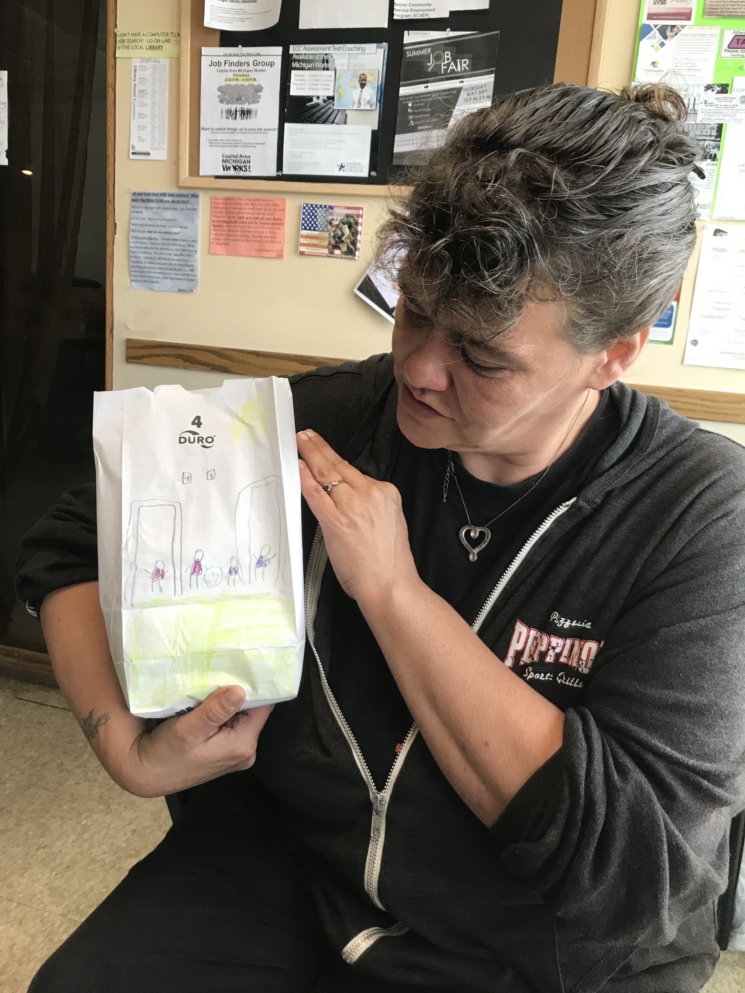 A woman with gray hair, wearing a black hoodie with a logo, holding a paper bag with a child's drawing attached. The drawing appears to depict four figures on a green field with two large structures and the sun. She is looking at the drawing with a g