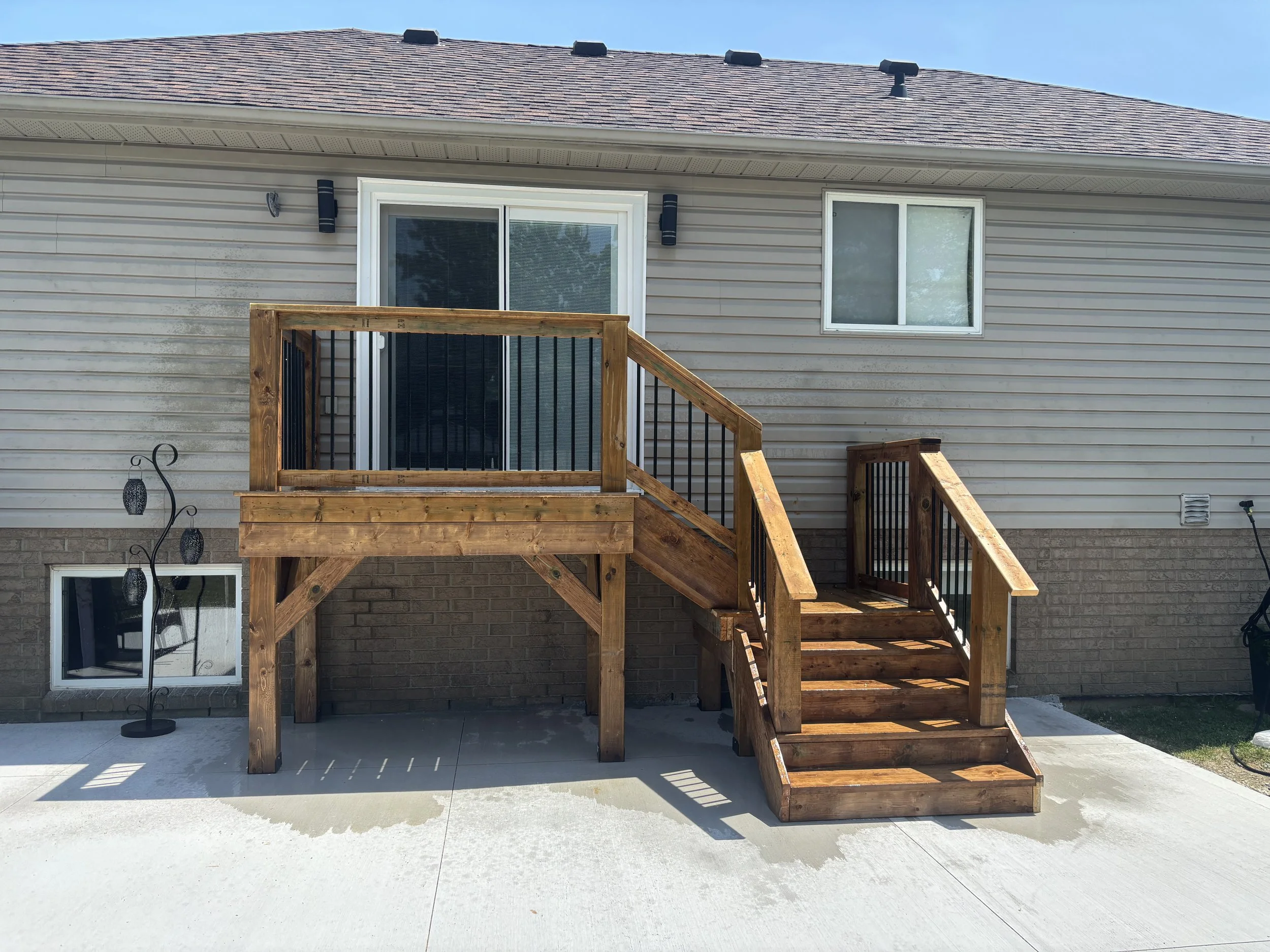 New wooden deck with stairs attached to the back of a beige house with two windows and sliding glass door, on concrete patio.