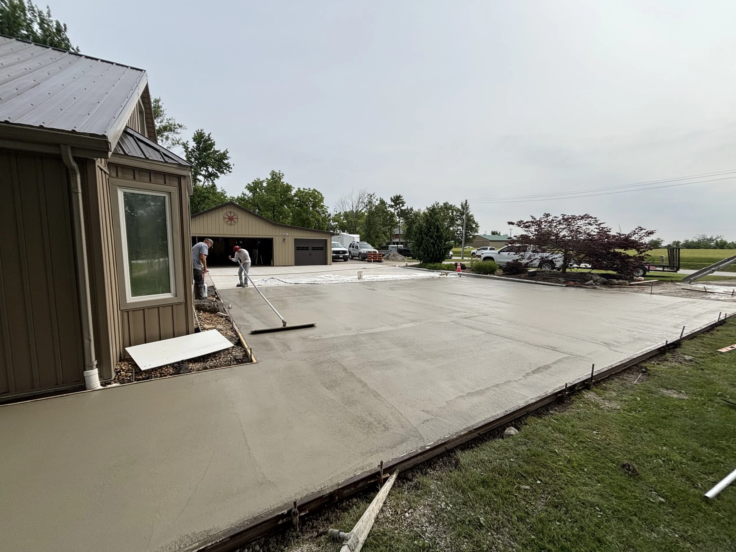 Two workers smoothing freshly poured concrete for a driveway in a residential area, with a house, garage, trees, and vehicles in the background.