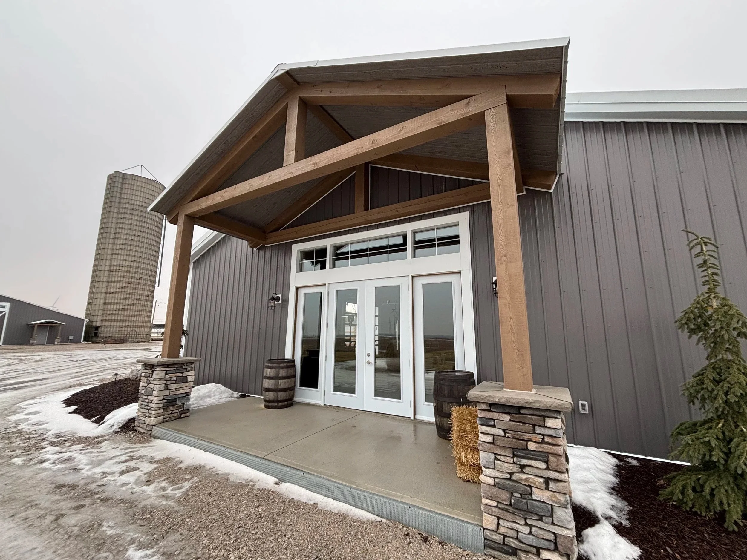 Exterior of a modern building with a stone porch, double glass doors, a small covered porch with wooden beams, barrels as planters, and snow on the ground.