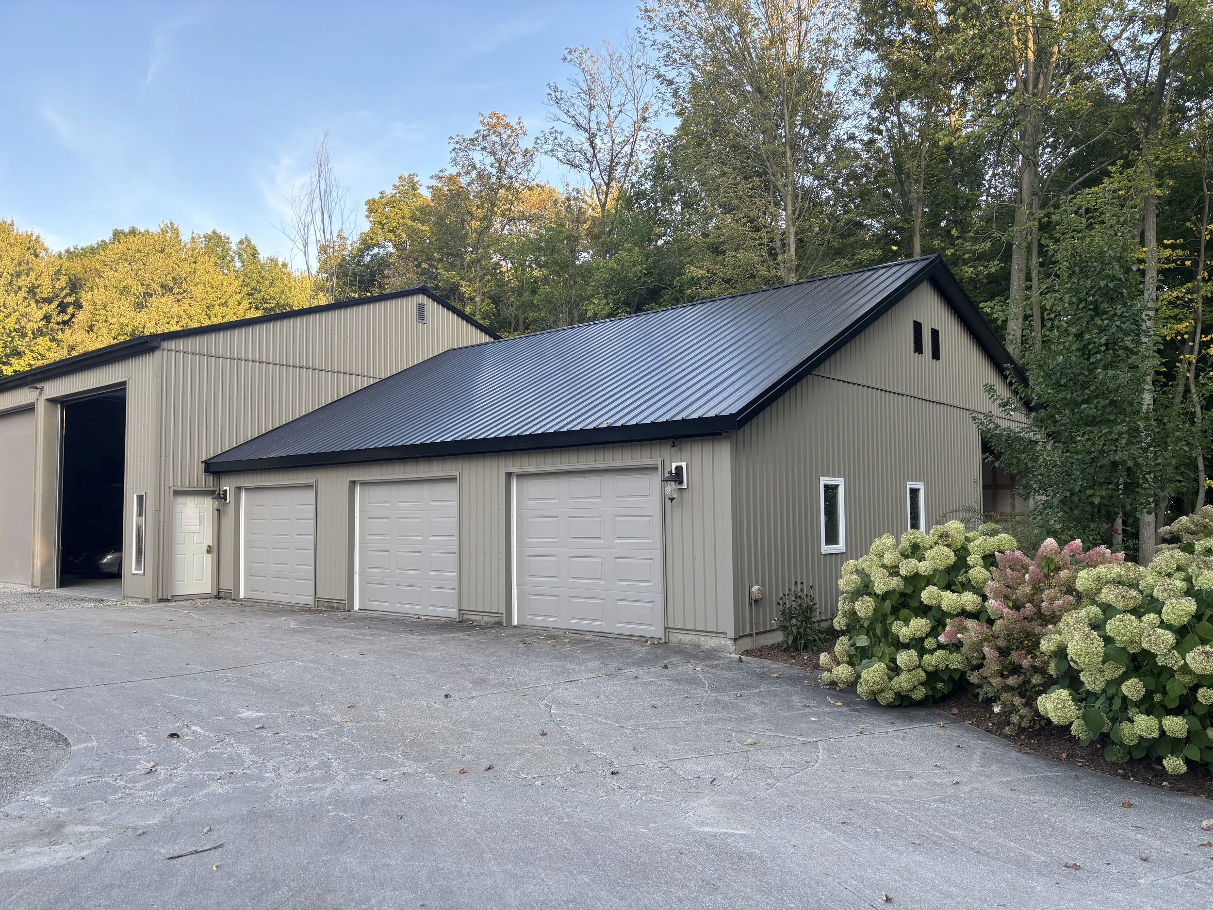 A beige metal building with three garage doors and a side entrance, surrounded by trees and bushes, under a partly cloudy sky.