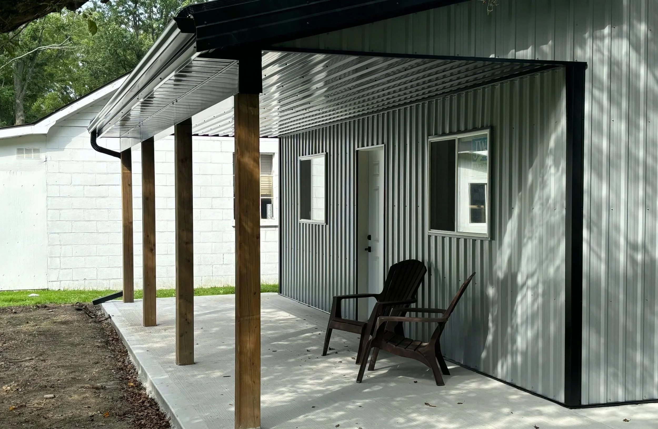 Back patio with two wooden chairs under a metal roofed awning adjacent to a metal building with two windows and a door, with a white brick building behind and trees visible.