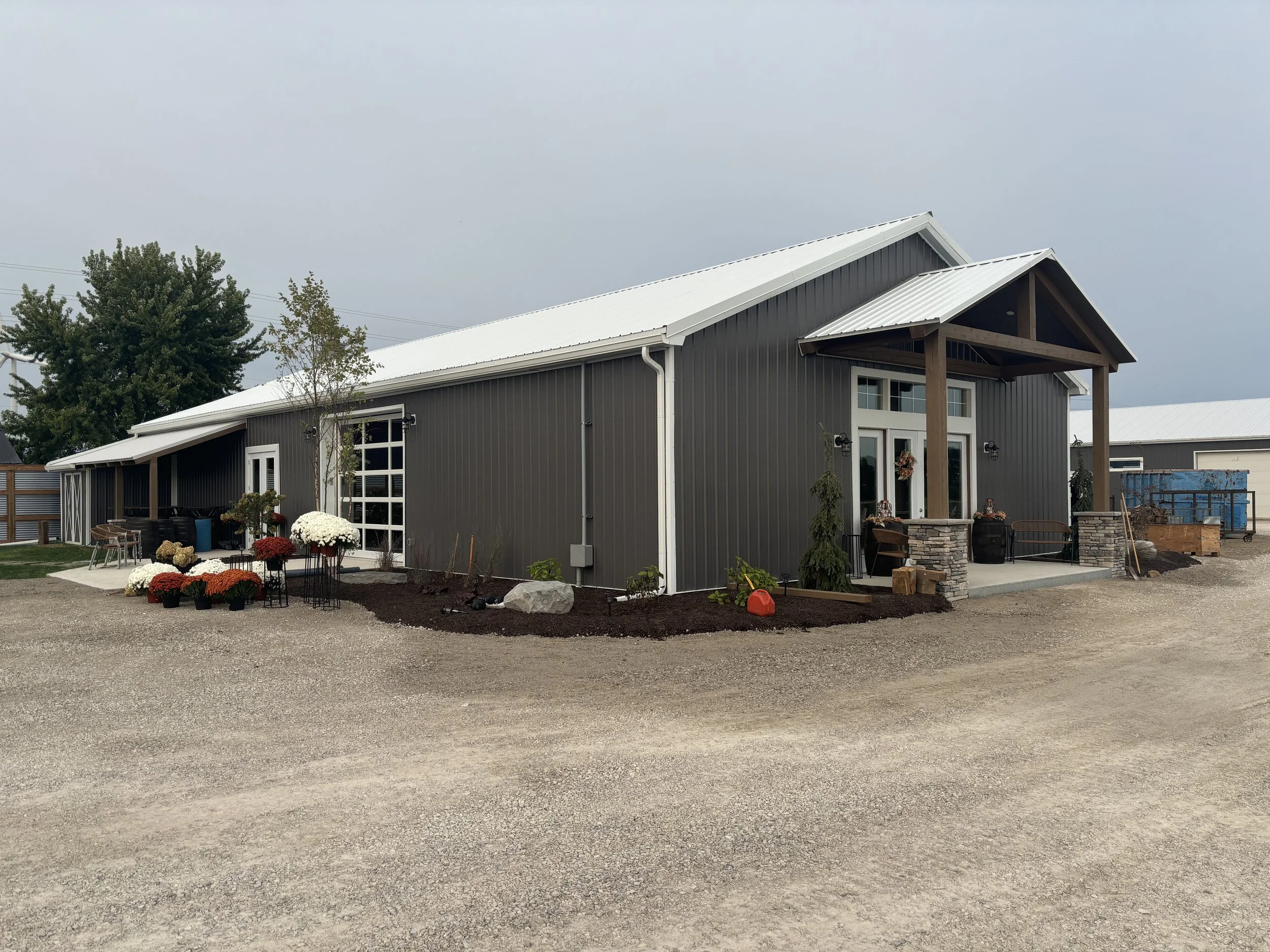 A modern barn-style building with dark gray metal siding, white trim, and a white metal roof. The front features a covered porch with wooden support beams, a door decorated with a wreath, and surrounding landscape with potted plants and flowers.