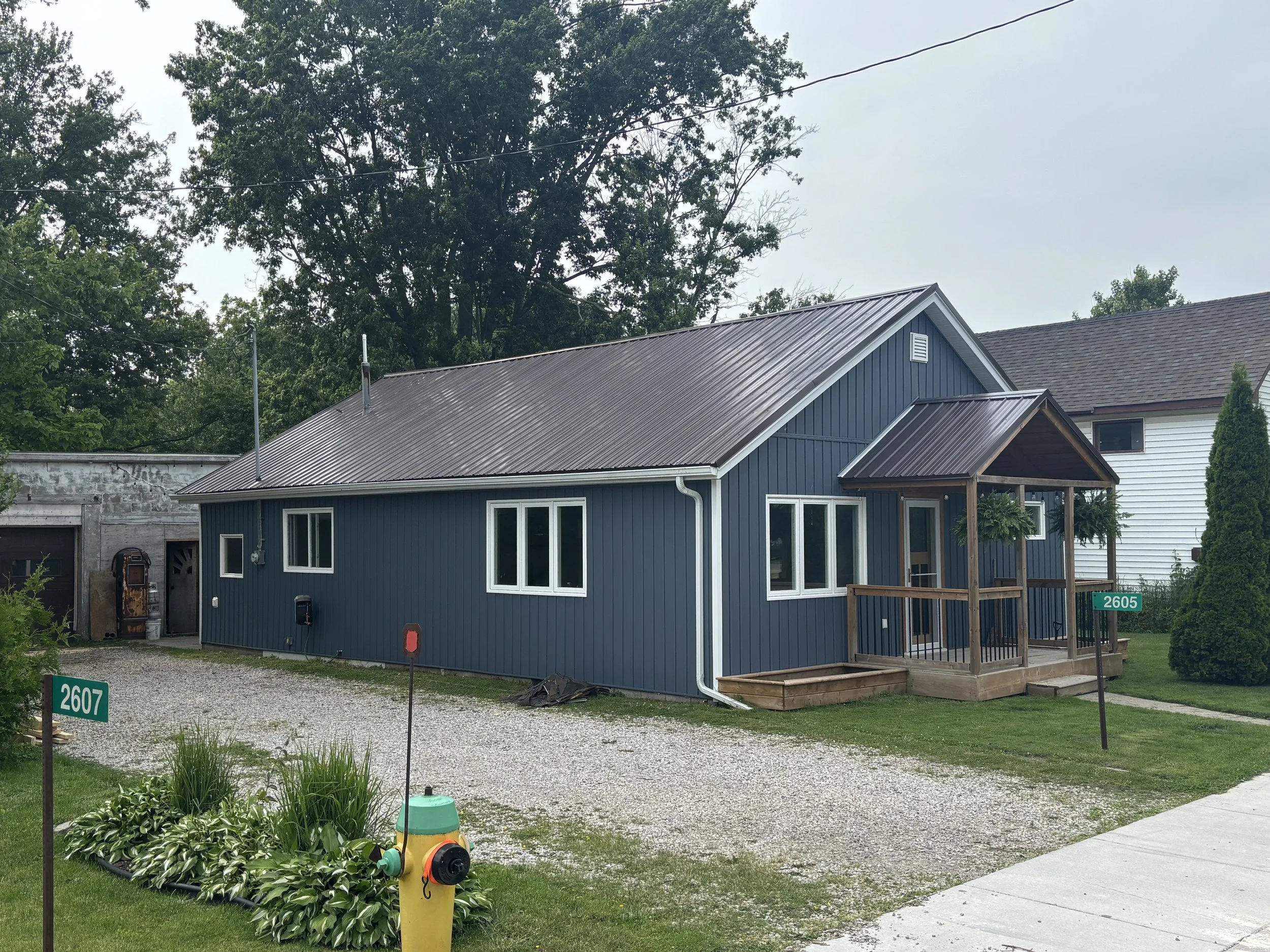 A blue house with a small porch, surrounded by grass and plants, with neighboring house and a gravel driveway.