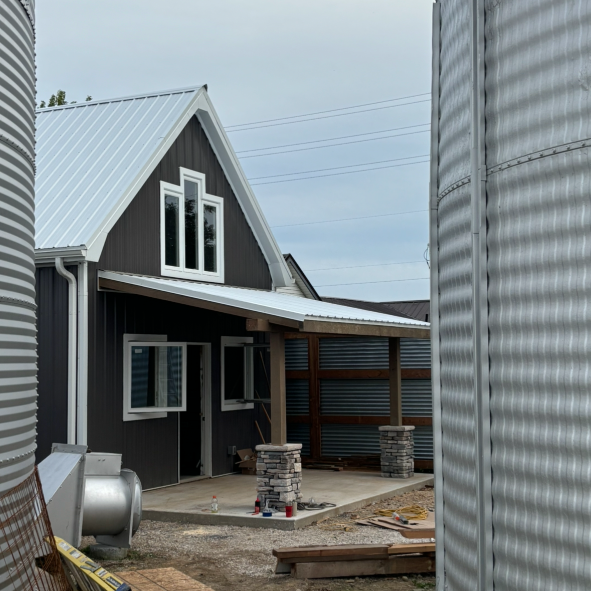 Construction site of a modern black house with metal roof and a small porch, surrounded by metal storage tanks.