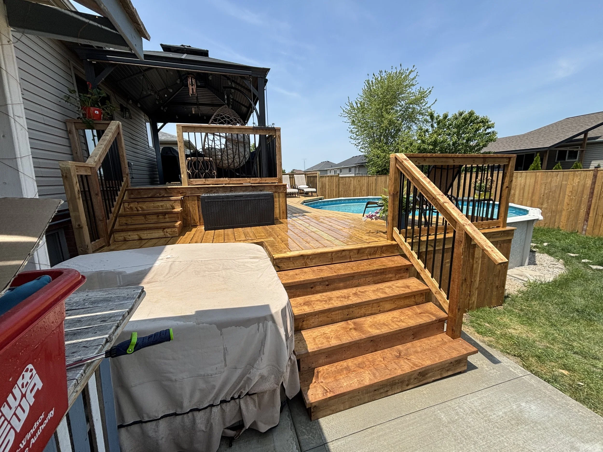 Backyard with a wooden deck, swimming pool, and a hot tub, surrounded by a wooden fence. Some outdoor furniture is visible, and the sky is clear and sunny.