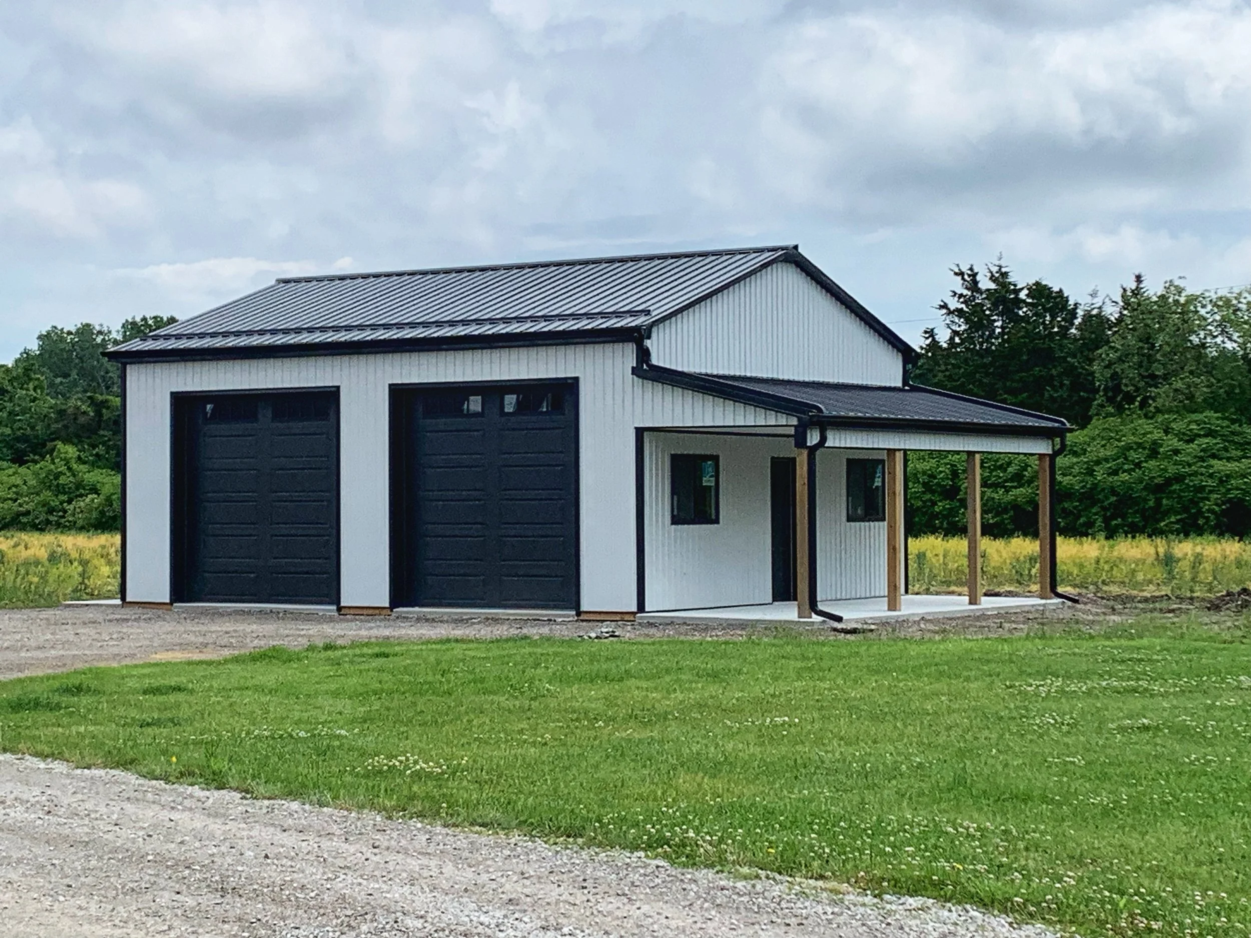 A newly constructed garage with two black doors, a white exterior, a metal roof, a small covered entrance with wooden support beams, and a gravel driveway surrounded by green grass and trees.