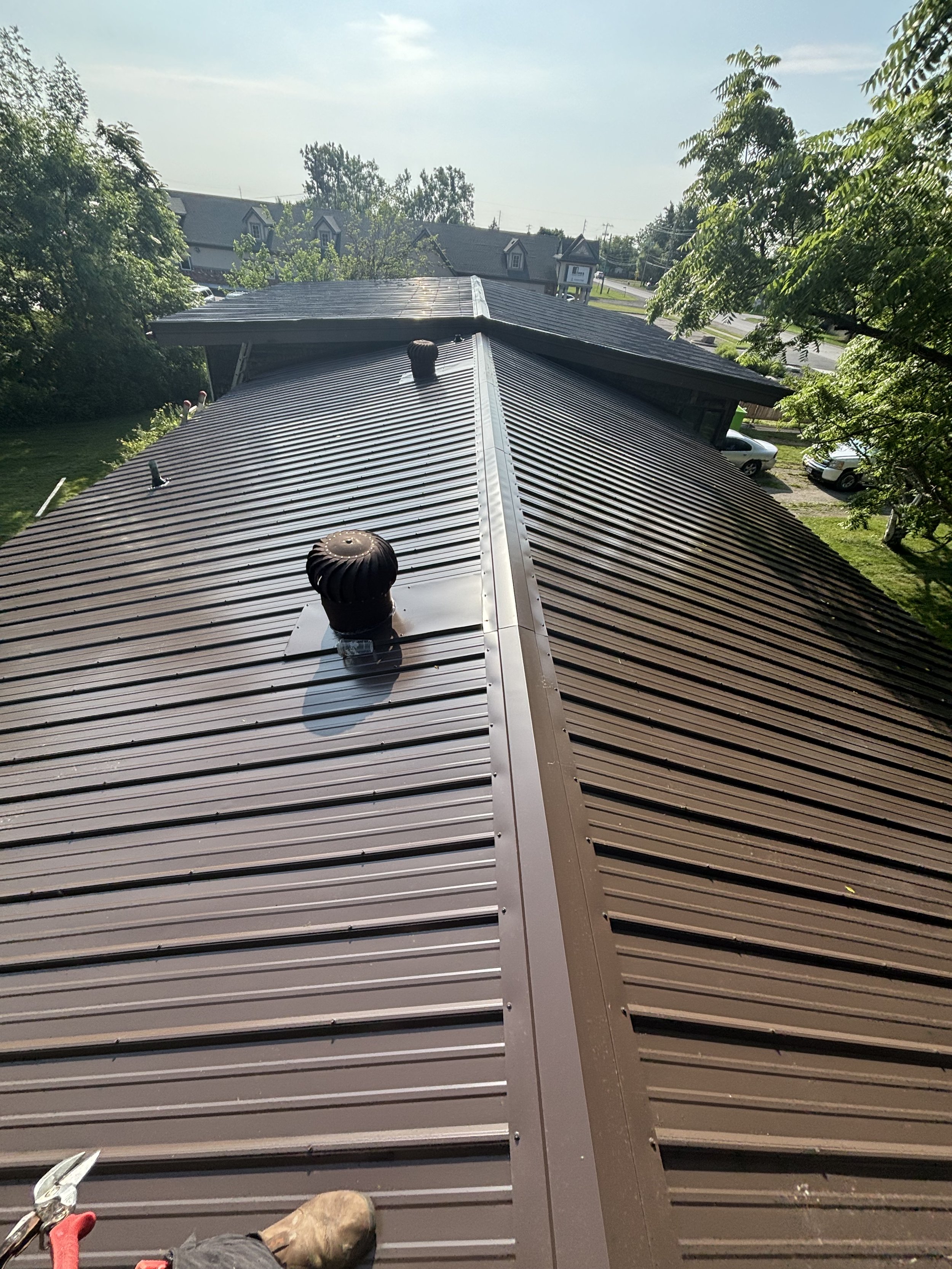 View of a metal roofing on a house, showing vents and chimney pipes, with trees and neighboring houses in the background.