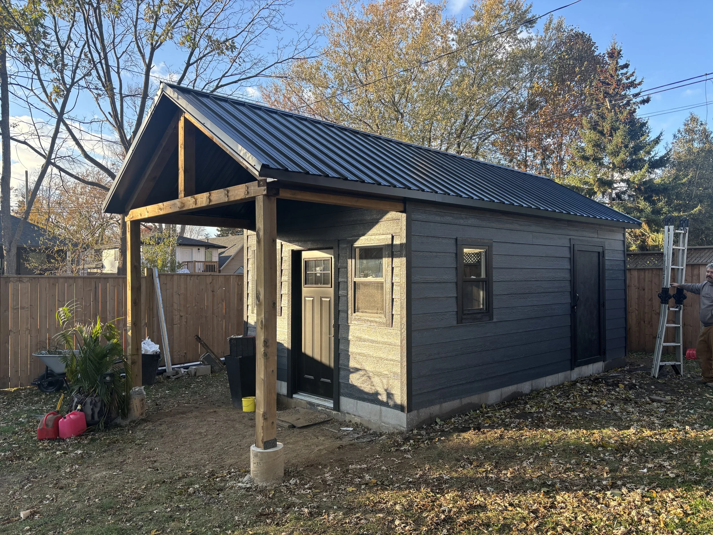 A small shed under construction with black siding and a metal roof, supported by wooden posts, situated in a backyard with a wooden fence, trees, and a man on a ladder to the right.