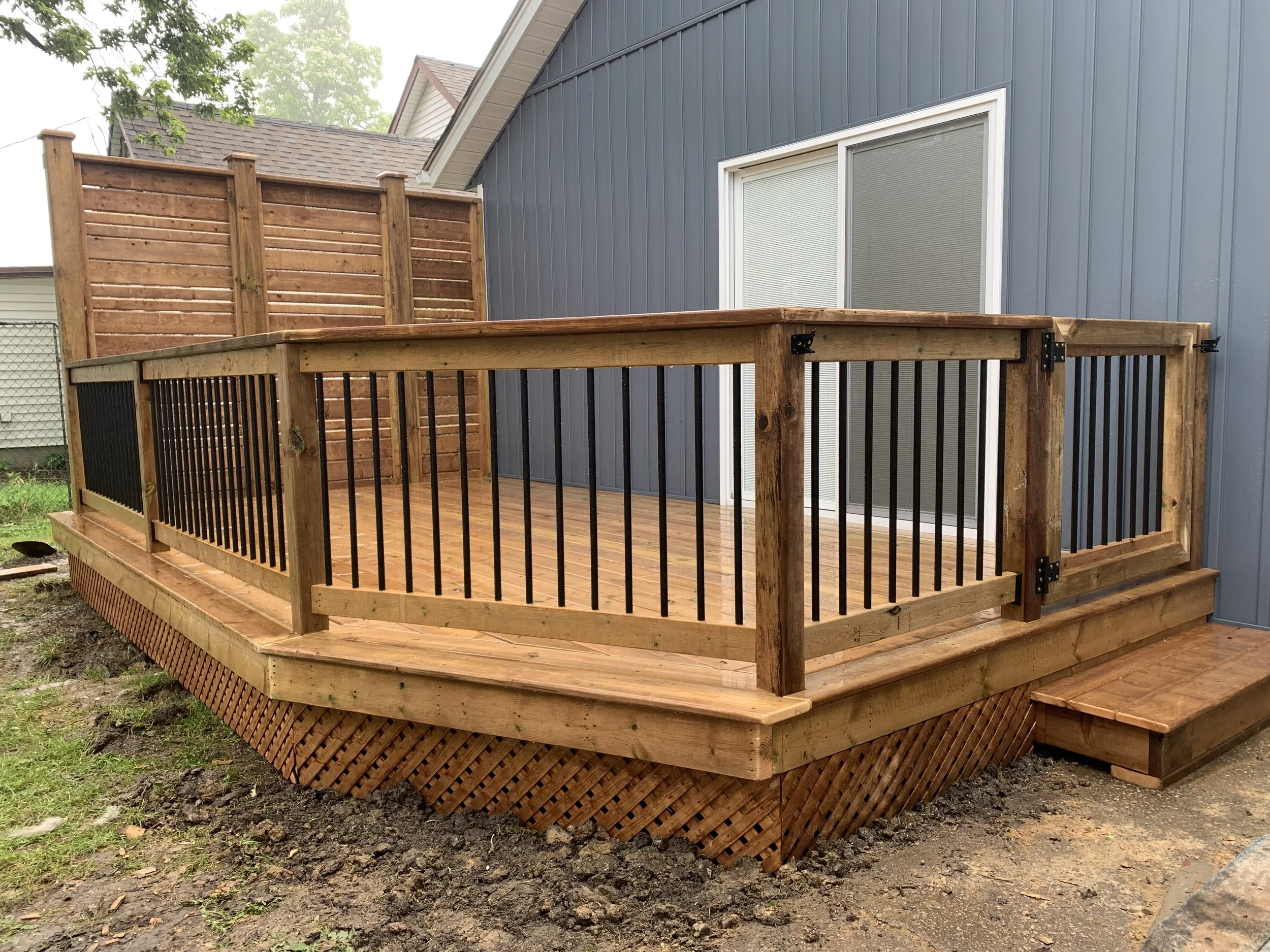 New wooden deck with black railings attached to a blue house with sliding glass door and steps leading up to the deck