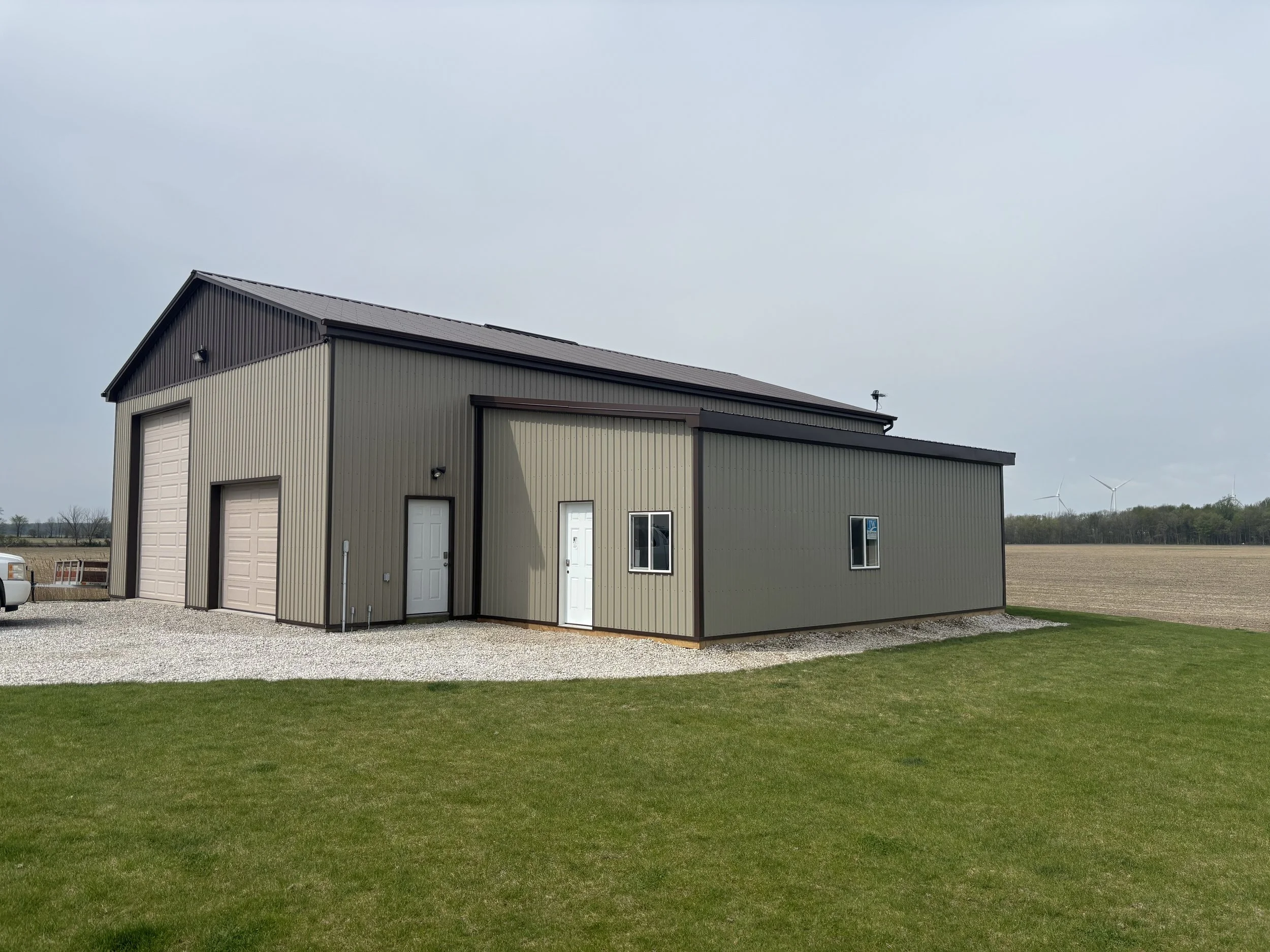 A modern metal building with a beige and dark brown exterior, featuring a garage door, a white pedestrian door, and small windows, situated on a grassy lawn with a gravel pathway, in a rural setting with open fields and wind turbines in the background.
