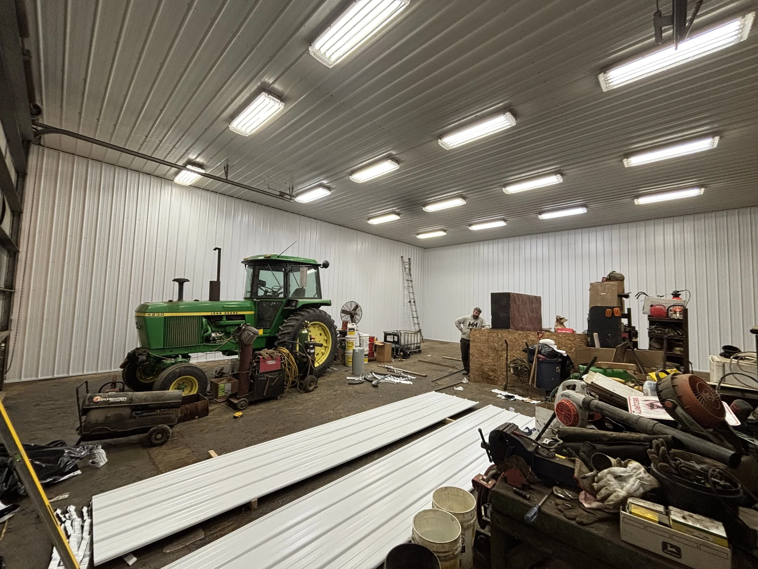 Interior of a workshop with a green John Deere tractor, tools, supplies, and two people working among construction materials and equipment.