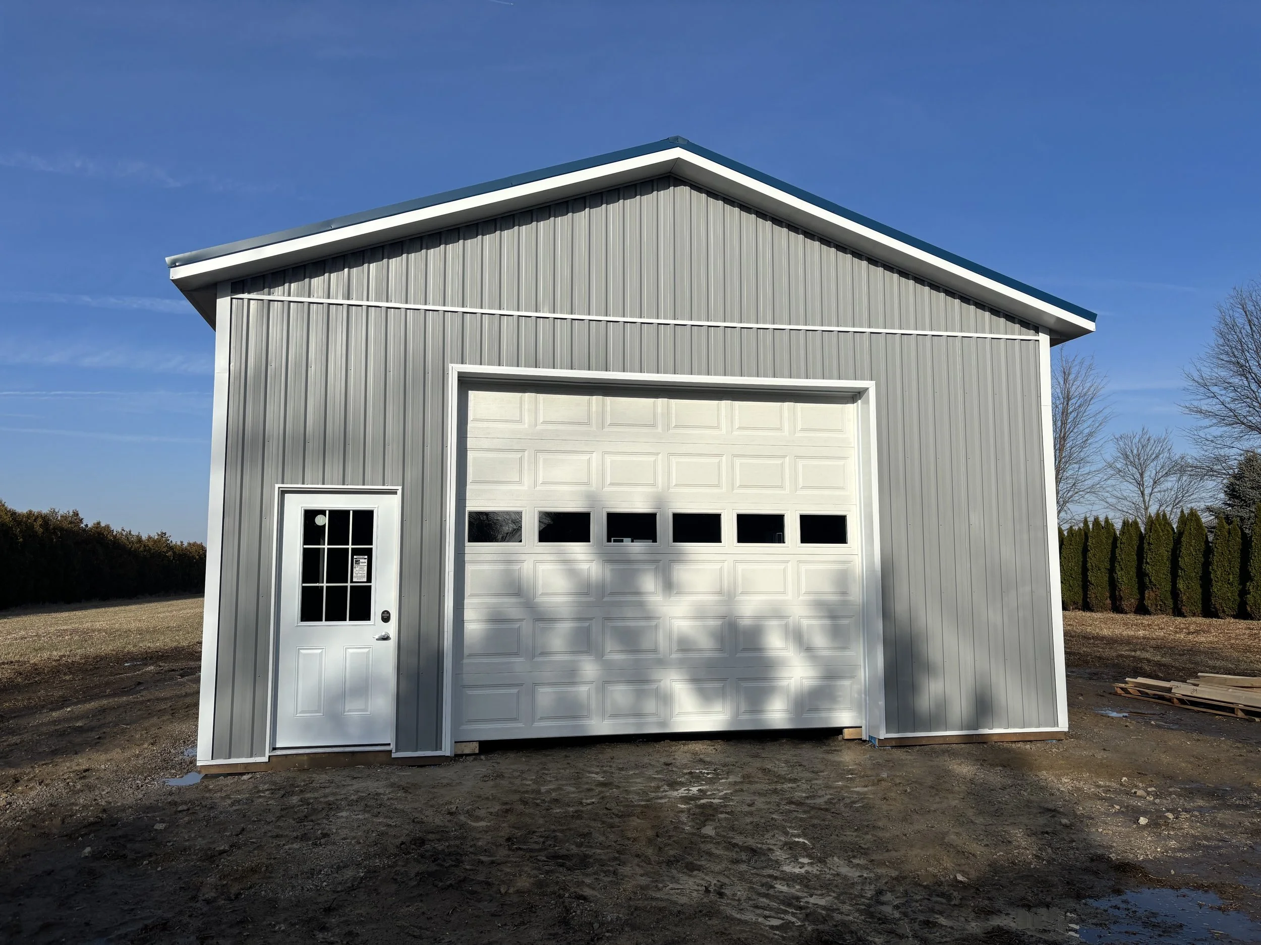 A gray metal garage with a white door and a small window, situated on a dirt lot under a clear blue sky.