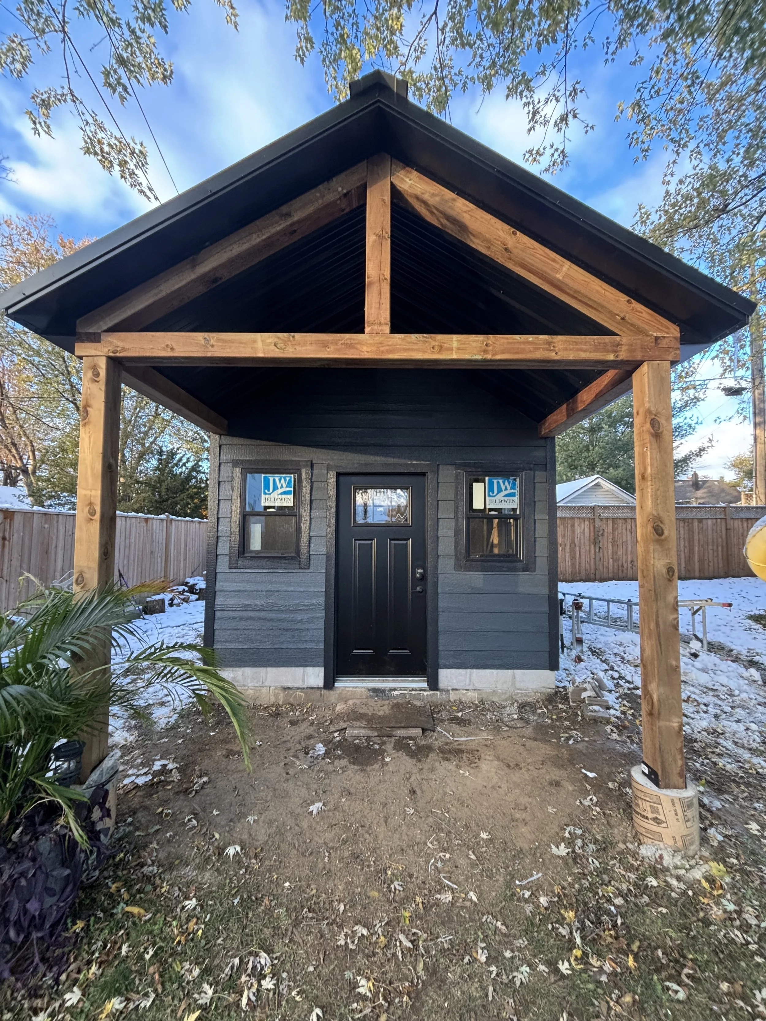 Small black house with a front porch under construction, with wooden posts supporting the roof, a black door, and two small windows, surrounded by a wooden fence and some snow on the ground.