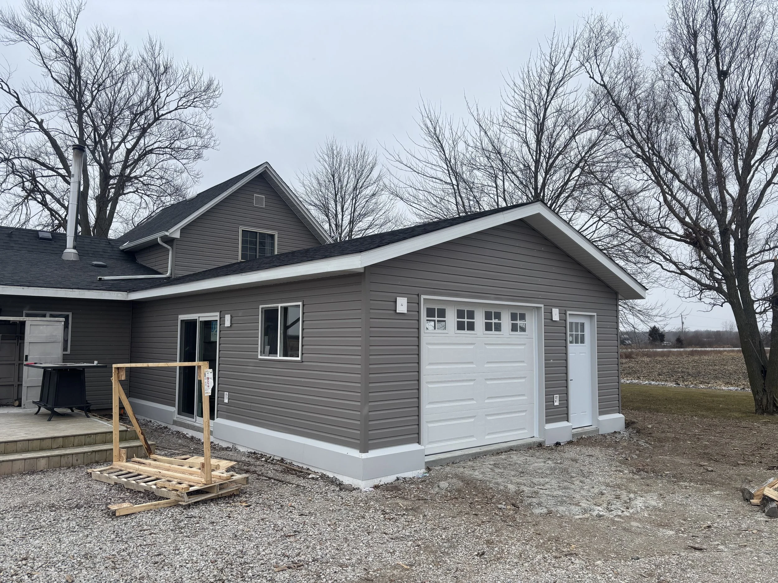 A newly constructed attached garage with gray siding and white doors, situated in a yard with bare trees and a cloudy sky.