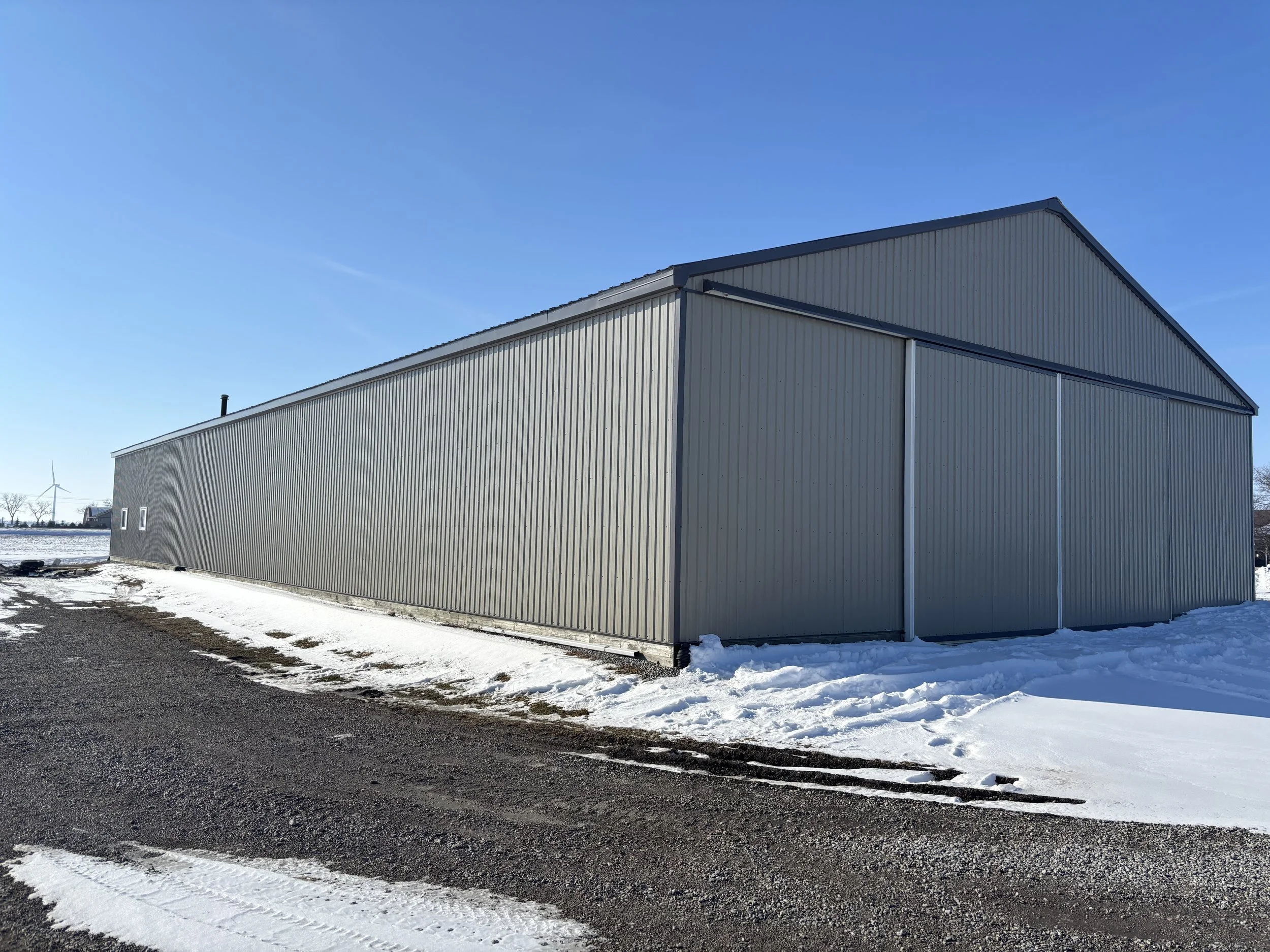 Large metal industrial building on snow-covered ground, with clear blue sky and wind turbine in the background.