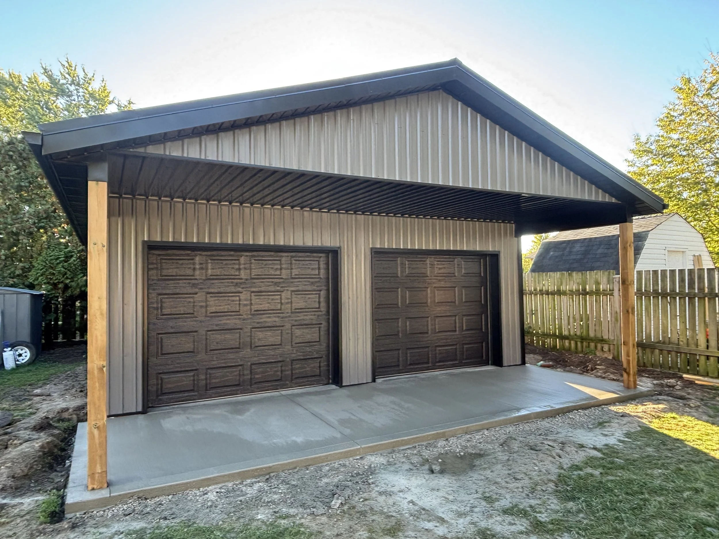 Newly constructed two-car garage with metal siding and dark brown garage doors, set on a concrete foundation with a dirt yard in front, surrounded by a wooden fence and trees.
