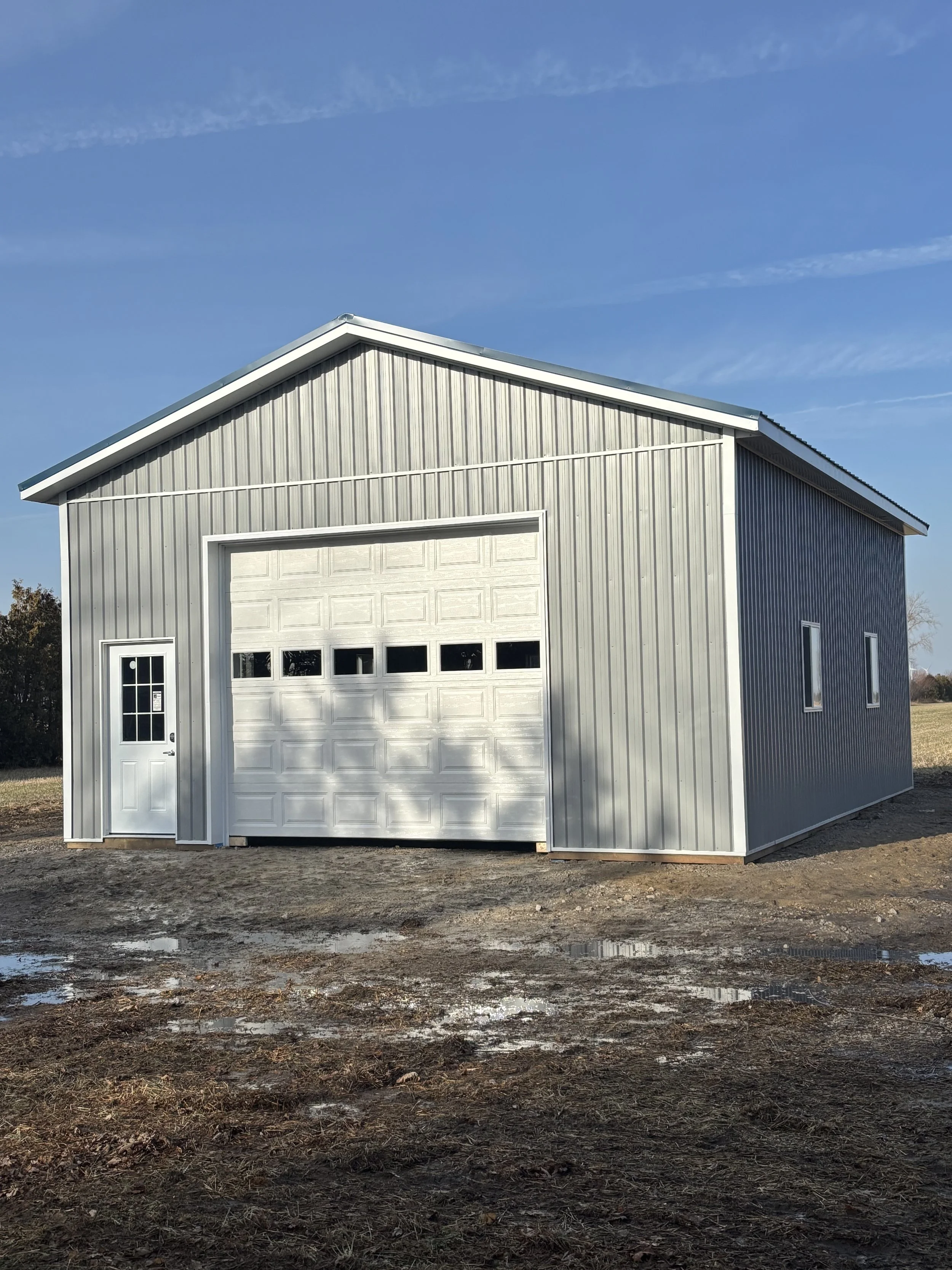 A metal building with a large garage door and a side door, situated on muddy ground with puddles, under a blue sky.