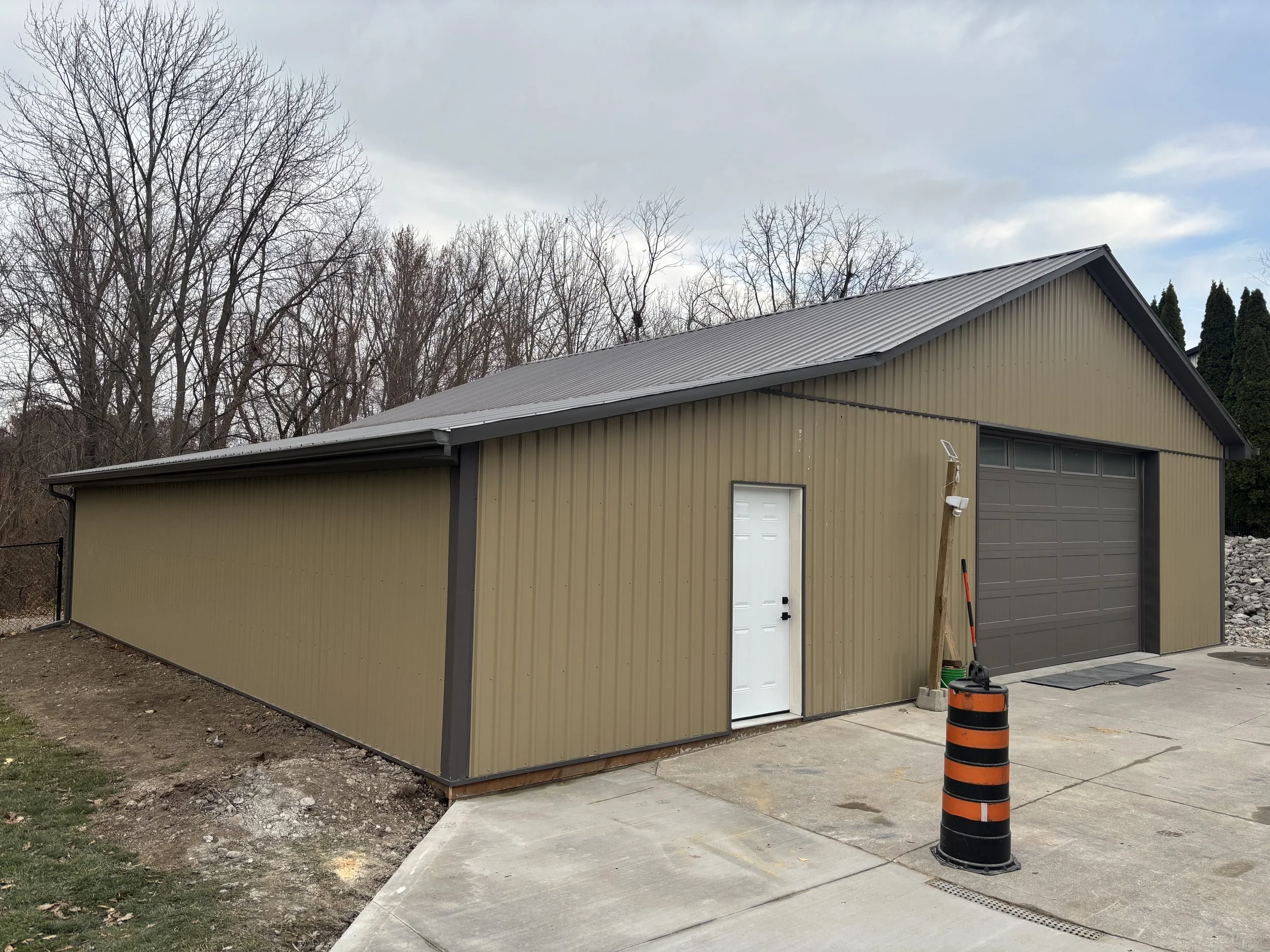 A beige metal building with a sloped roof, a white door, and a garage door, situated on a concrete driveway with a black and orange barrel in the foreground and leafless trees in the background.