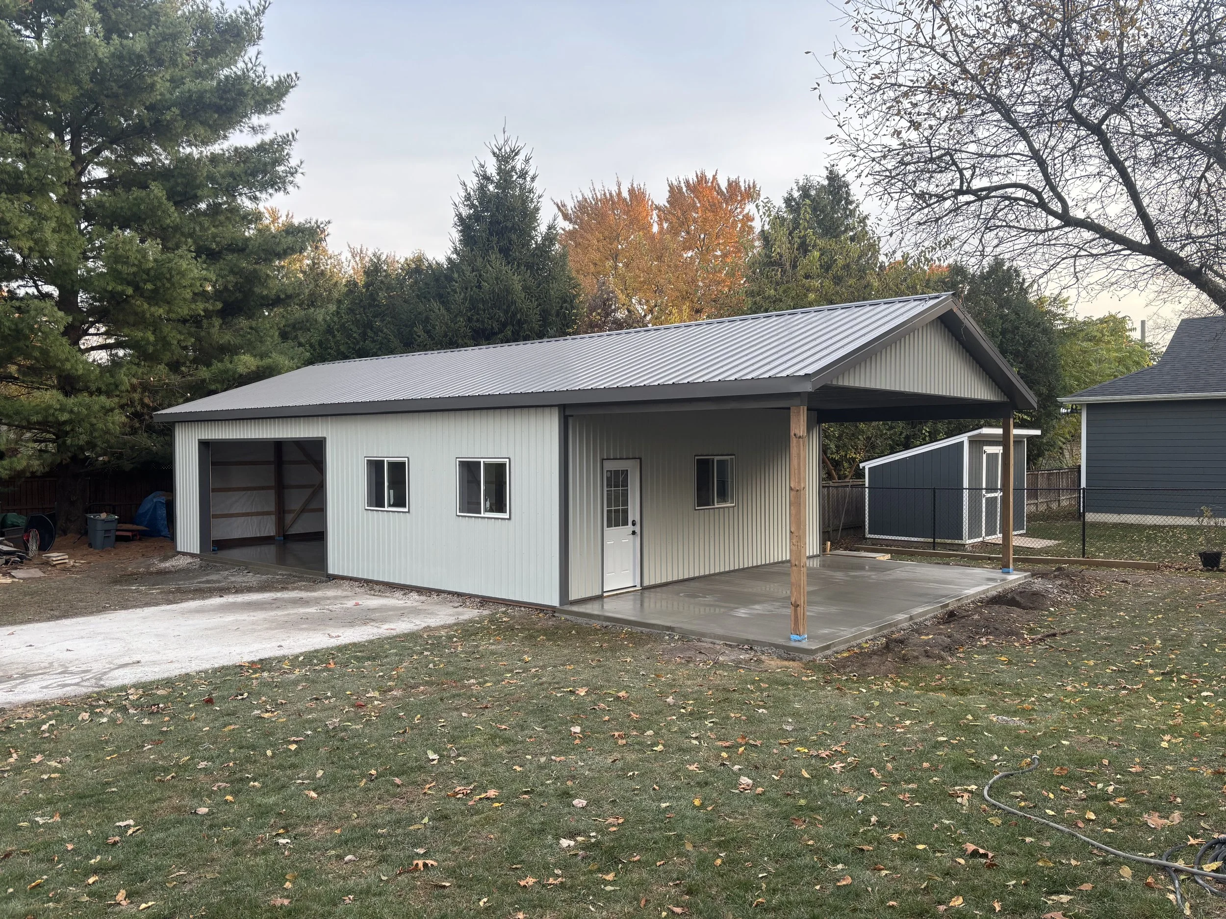 A newly constructed metal outbuilding with a sloped roof, two small windows, a door, and a covered concrete patio in a backyard with trees and grass.