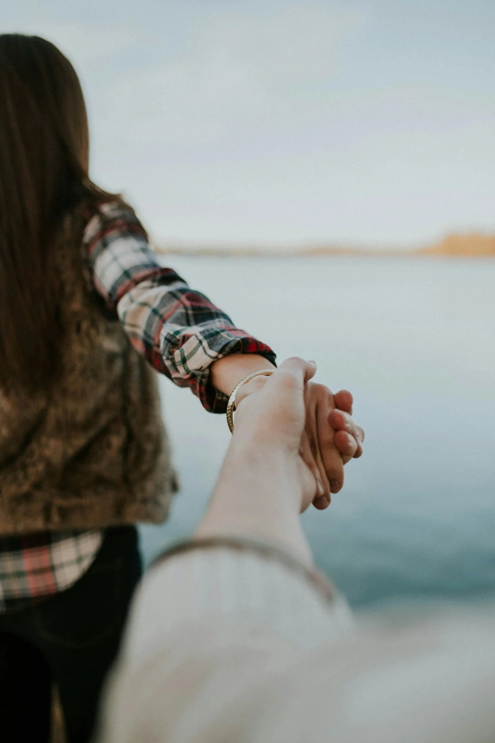 A person with long brown hair, wearing a plaid shirt, holding hands with a child against a blurred outdoor background.