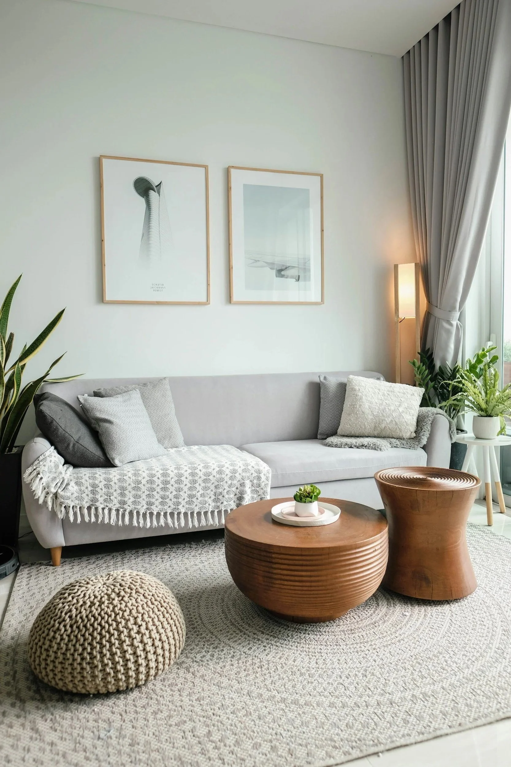A cozy living room with a gray sofa, several pillows, a textured blanket, modern framed art on the wall, potted plants, and wooden coffee tables on a patterned rug.
