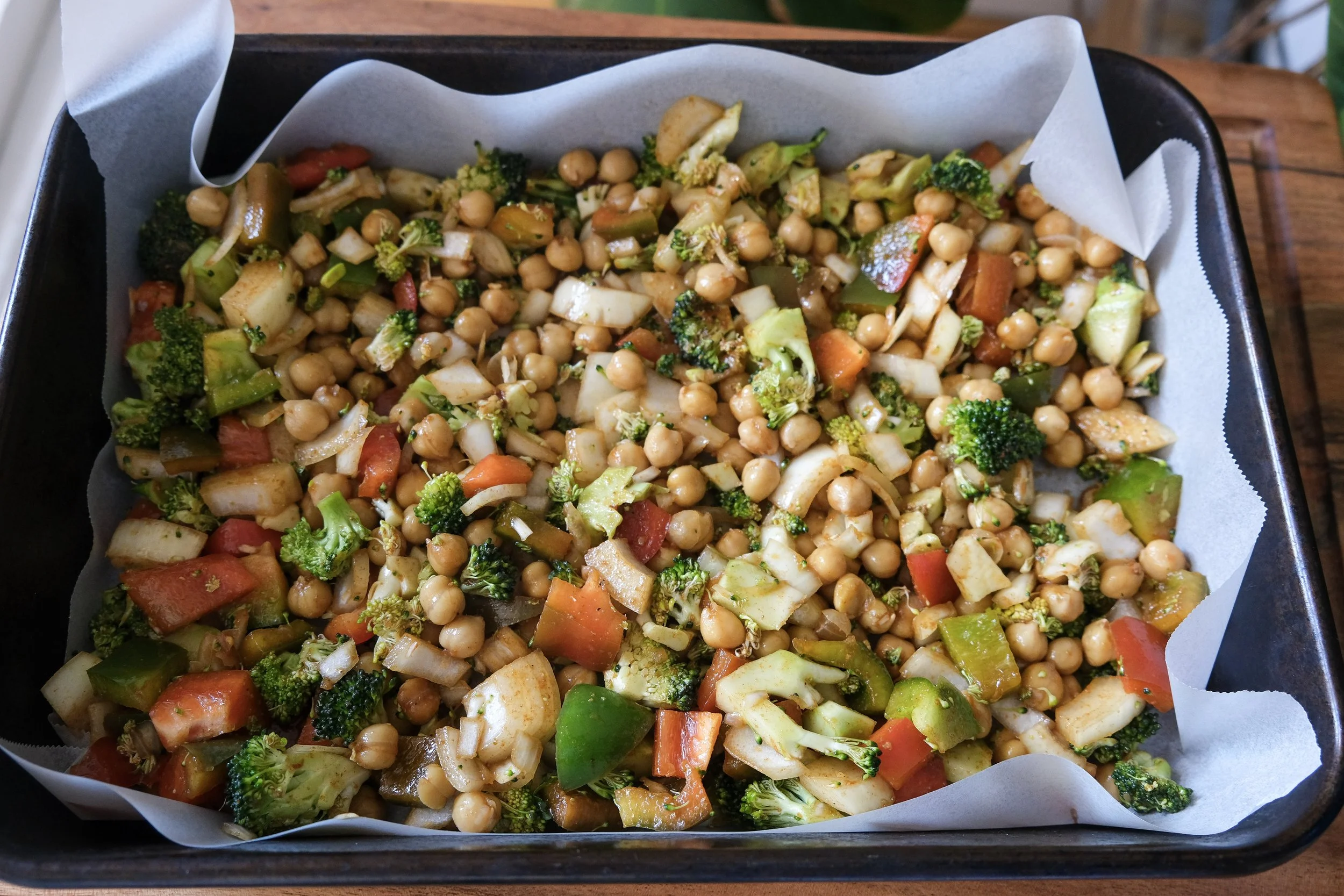 A baking sheet lined with parchment paper filled with seasoned chickpeas, broccoli florets, chopped onions, and diced bell peppers ready for roasting.