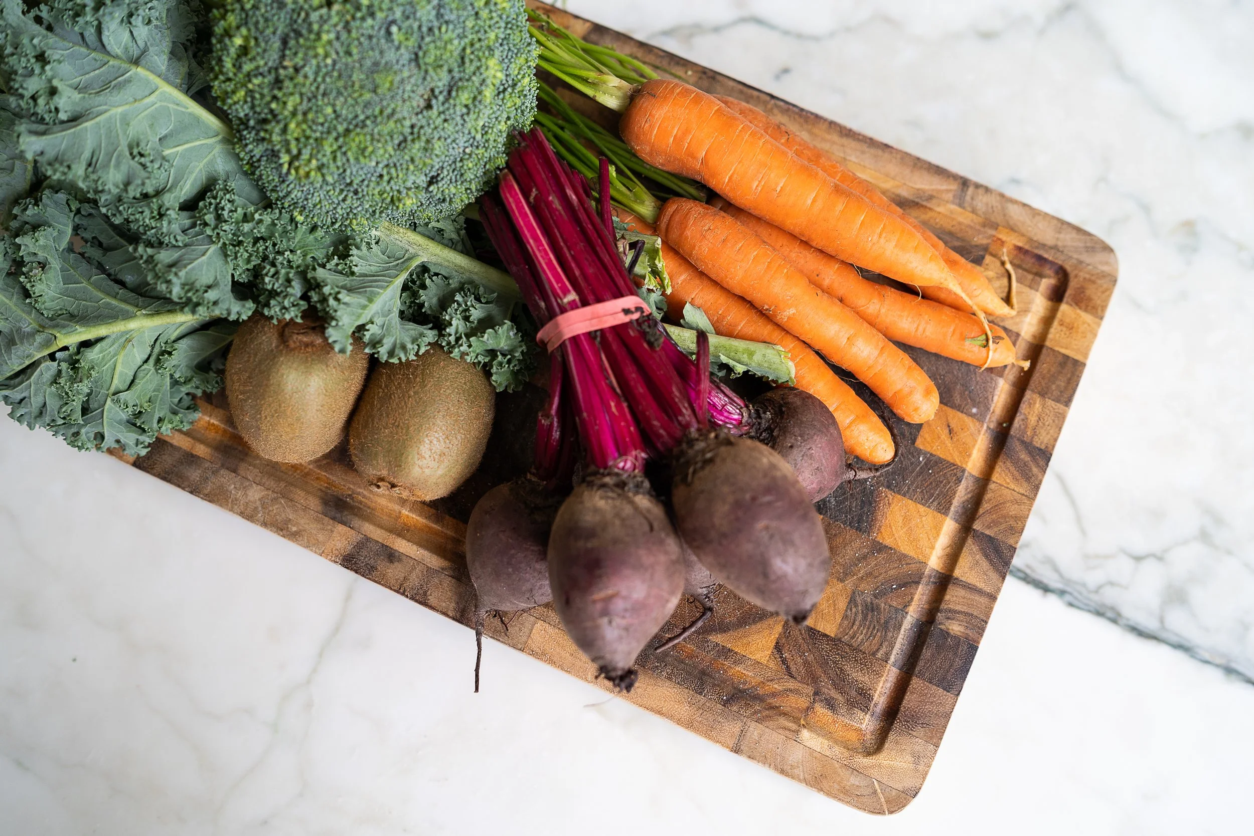 A wooden cutting board filled with fresh gut healthy foods including broccoli, kale, carrots, beets, and kiwi fruits on a marble countertop.
