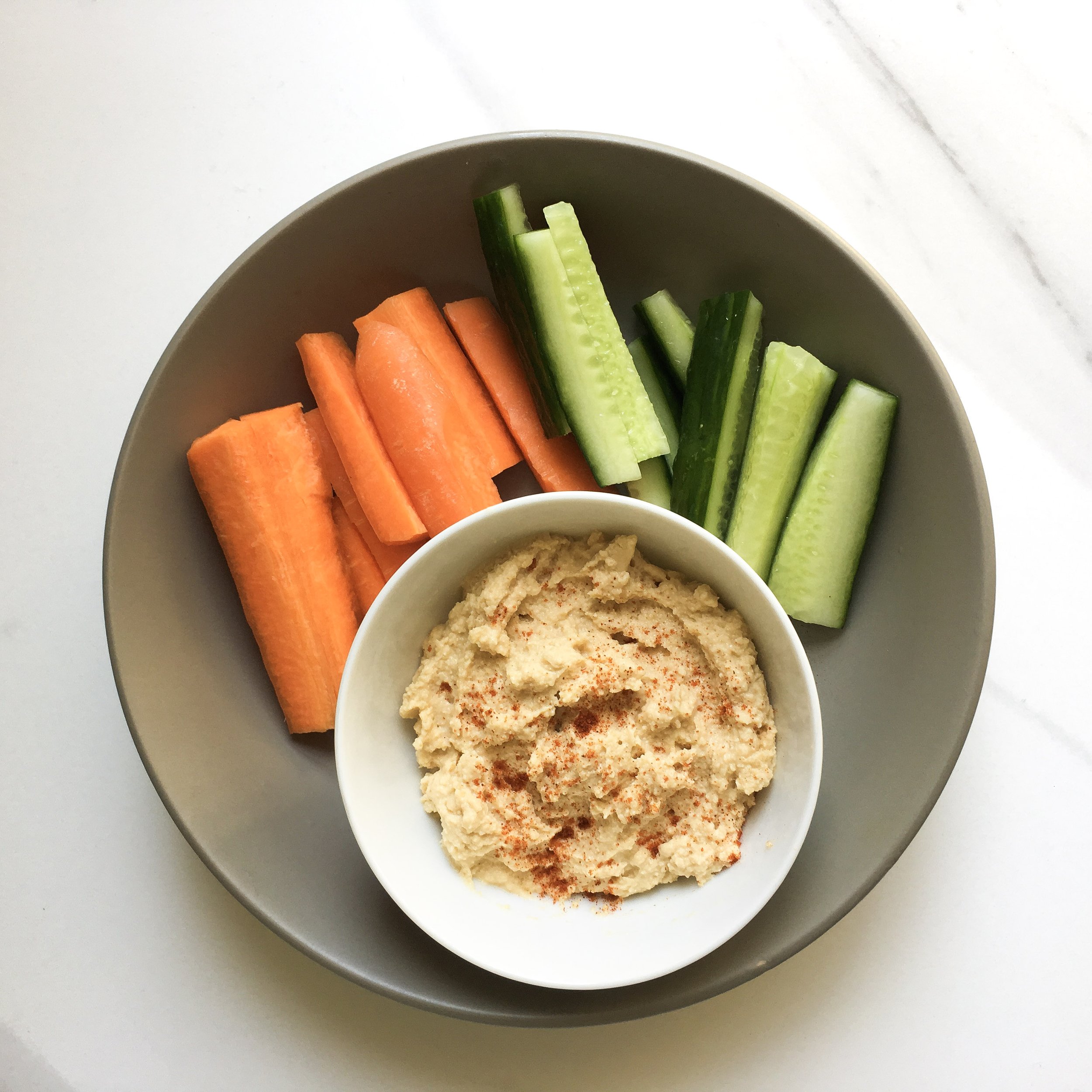 A top-down view of a small white bowl of homemade hummus sprinkled with paprika, served inside a grey platter with fresh carrot sticks and cucumber spears on a marble background.