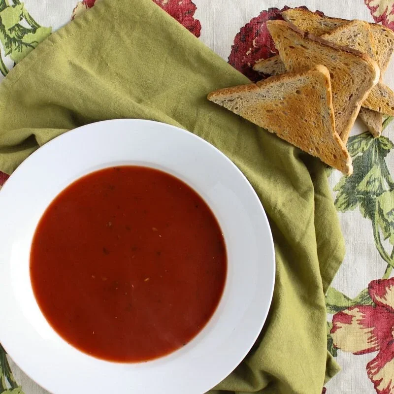 A white bowl of smooth low FODMAP tomato soup served with triangles of toasted gluten-free bread on a green linen cloth.