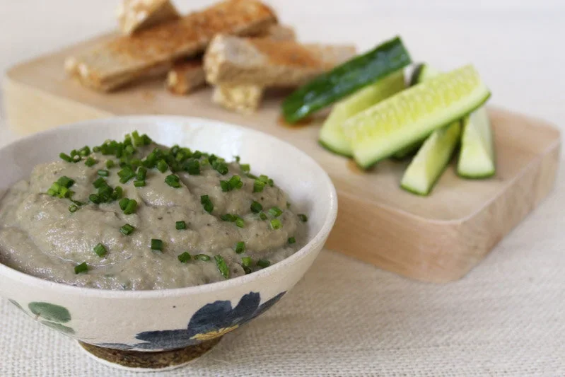 A bowl of creamy roasted eggplant dip garnished with fresh chives, served with cucumber sticks and toasted bread.