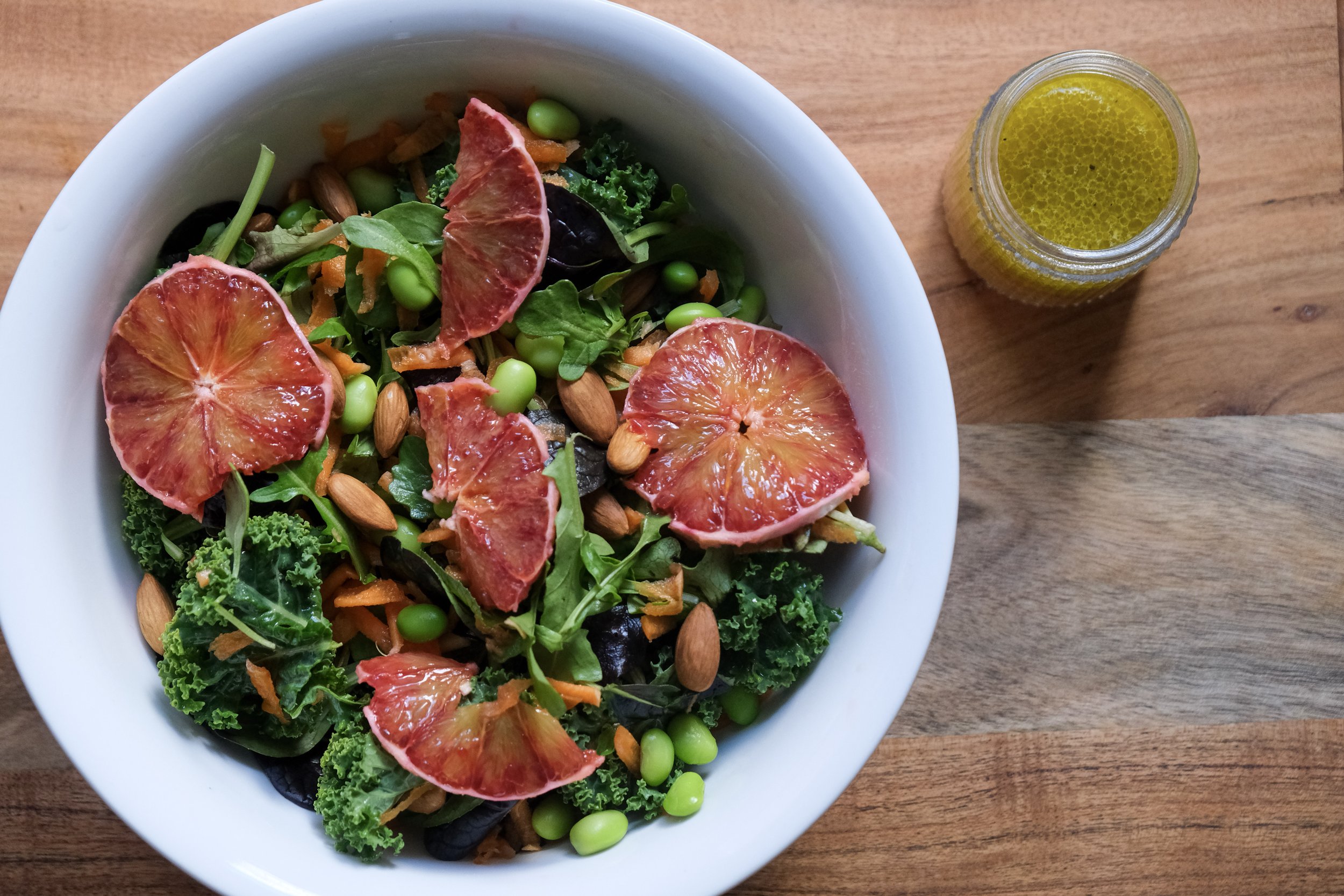 A top-down view of a white bowl filled with a kale and edamame salad topped with blood orange slices and whole almonds, with a small jar of miso dressing on a wooden table.