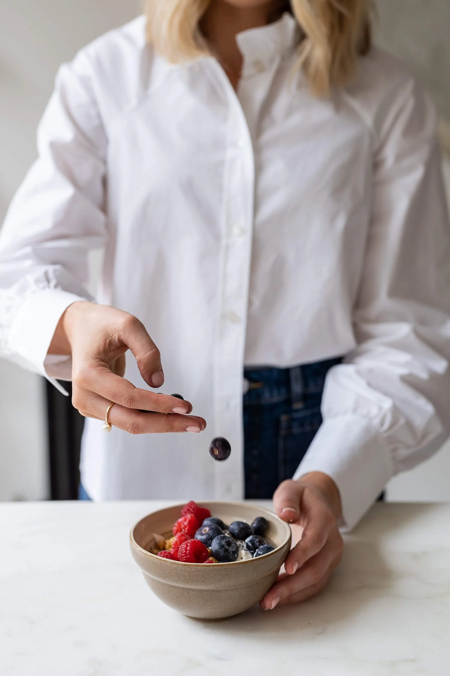 A woman in a white shirt is sprinkling blueberries and raspberries into a beige bowl on a white surface.