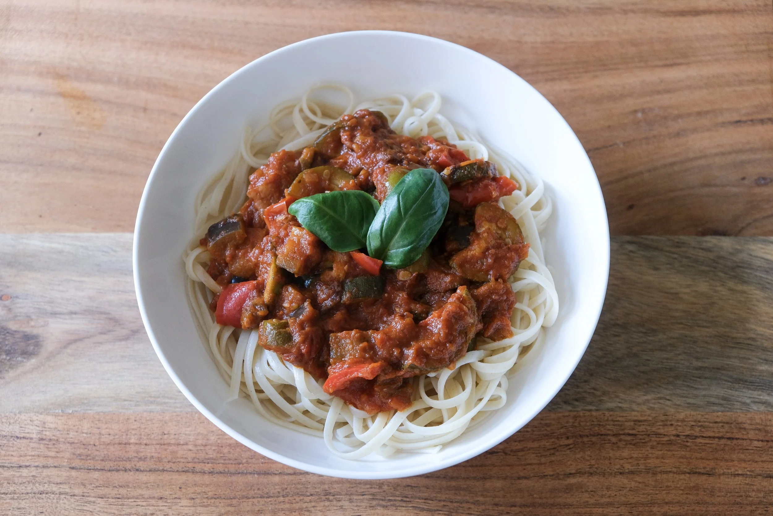 A white bowl of long pasta noodles topped with a rich tomato vegetable sauce containing roasted zucchini, eggplant, peppers, and fresh basil leaves.