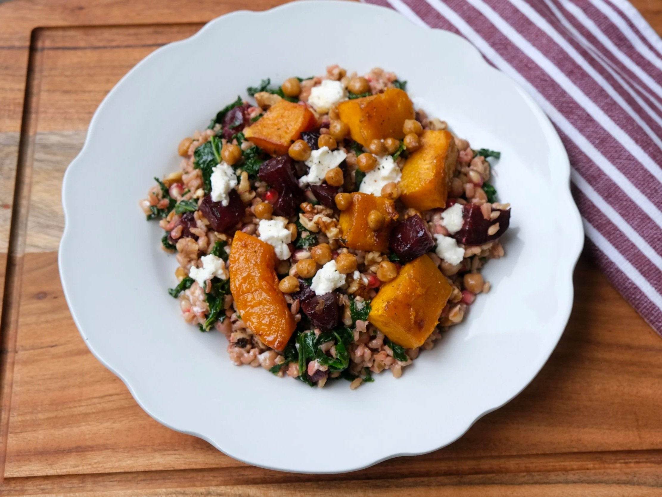 A white plate topped with a grain salad featuring roasted pumpkin wedges, diced beetroot, kale, crispy chickpeas, pearl barley, pomegranate seeds, and crumbled goat cheese.