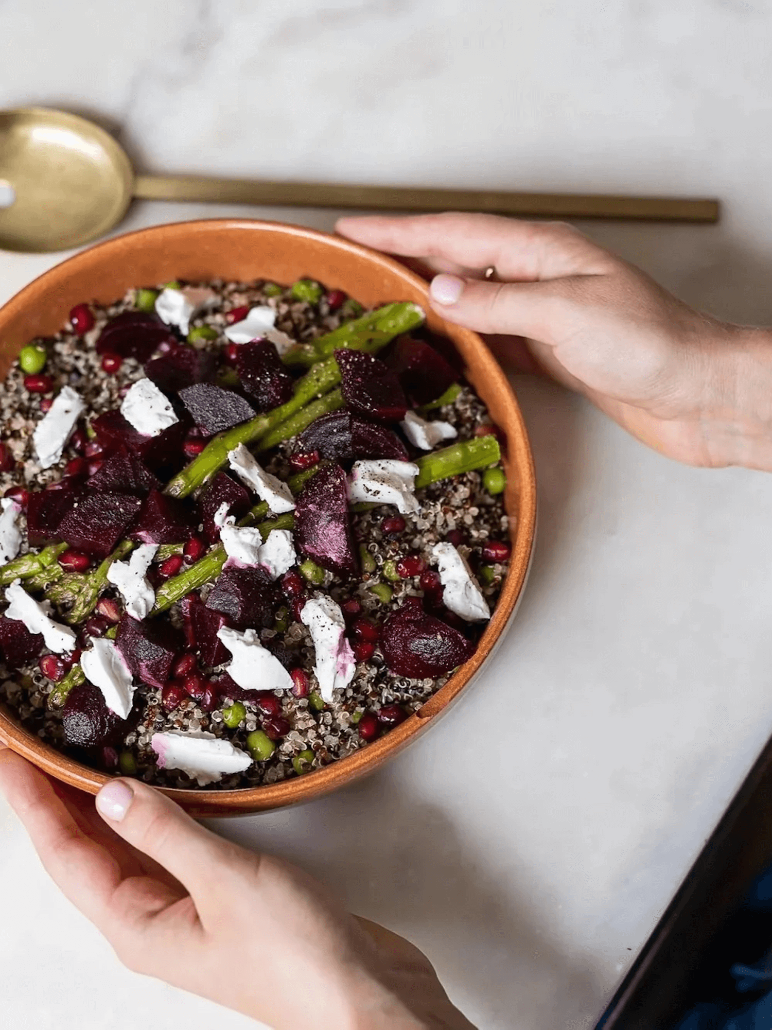 A person holding a bowl of colorful salad with beets, quinoa, green beans, pomegranate seeds, and crumbled cheese.