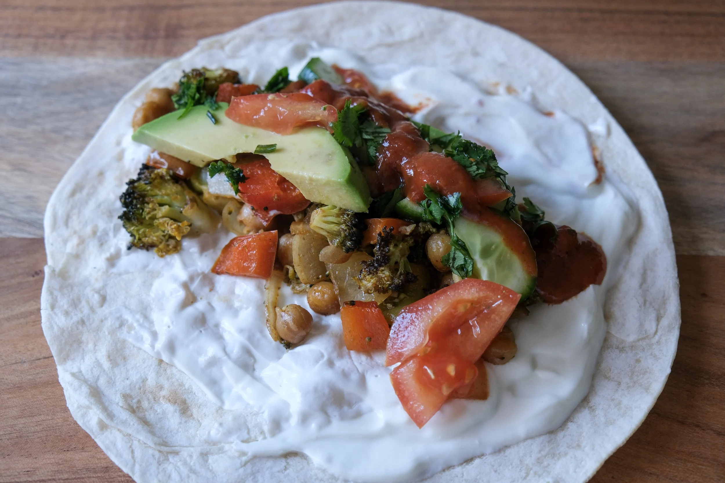An open flour tortilla spread with Greek yogurt and filled with roasted chickpeas, charred broccoli florets, diced tomatoes, sliced cucumber, avocado, salsa, and fresh herbs.