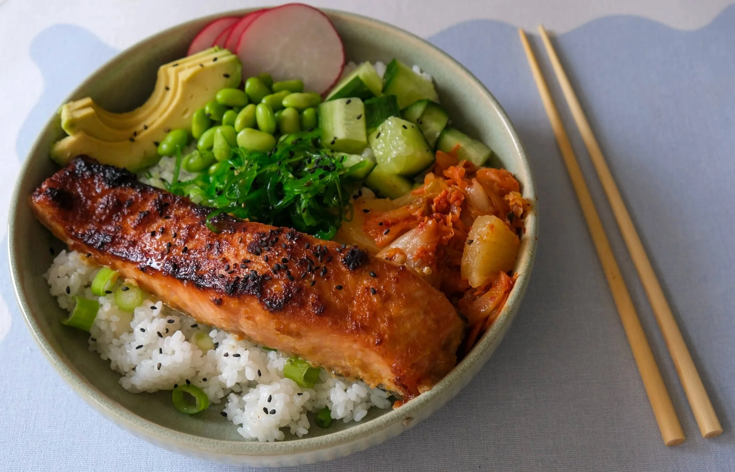 A glazed miso salmon fillet served over white rice in a bowl, surrounded by sliced avocado, edamame, diced cucumber, radishes, kimchi, and seaweed salad.