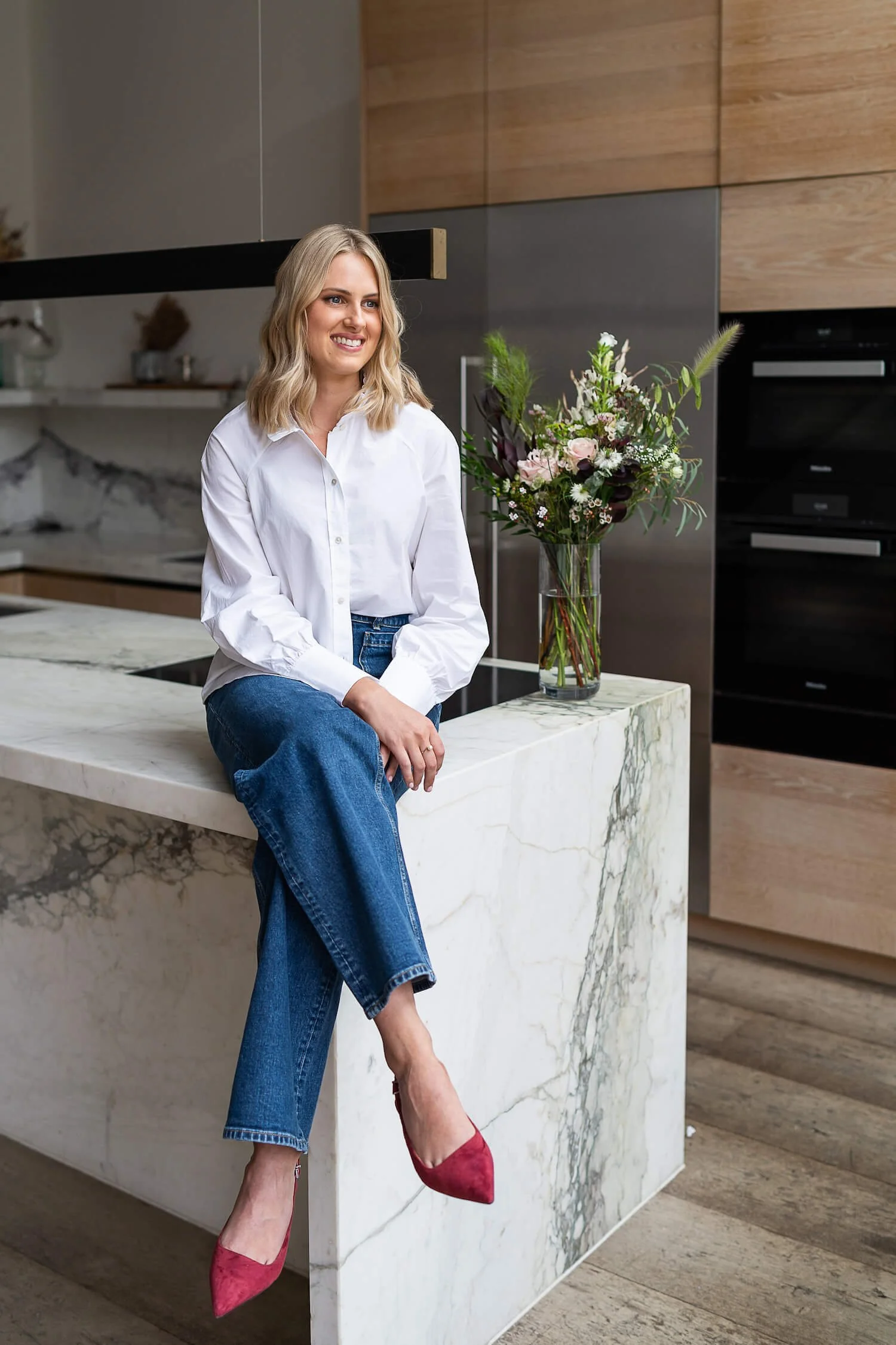 A woman sitting on a marble kitchen island with a bouquet of flowers nearby, in a modern kitchen with wooden cabinets and stainless steel appliances.