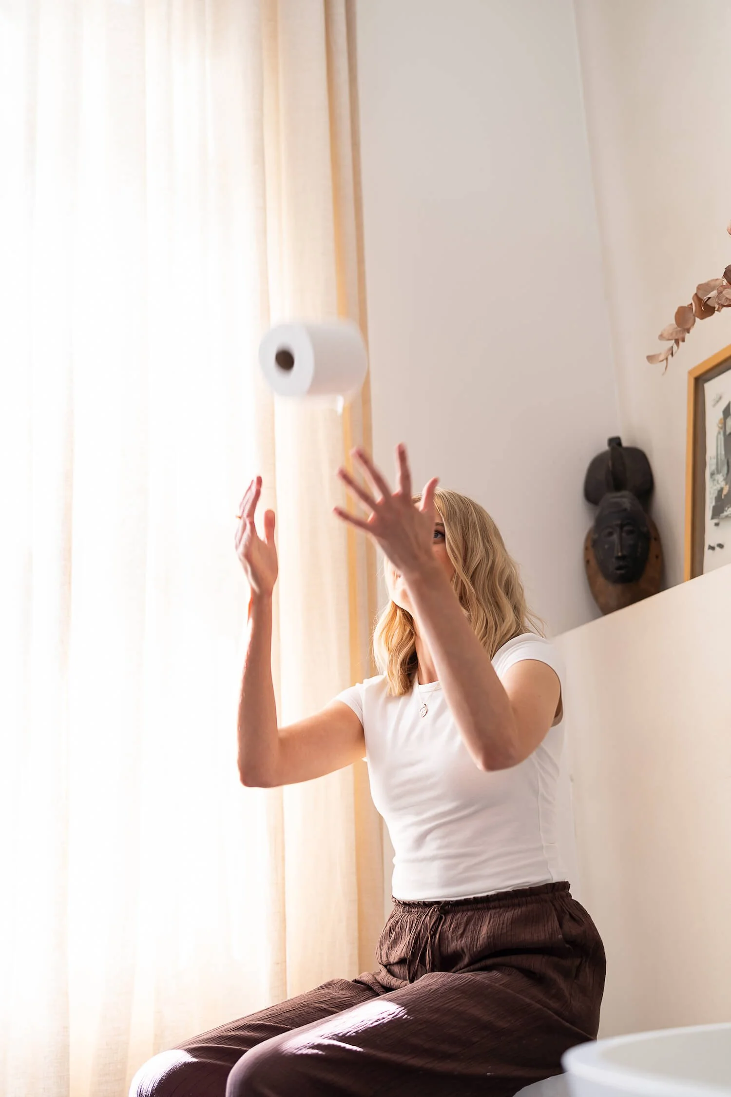 Woman sitting on a chair in a bright room, tossing a roll of toilet paper into the air.