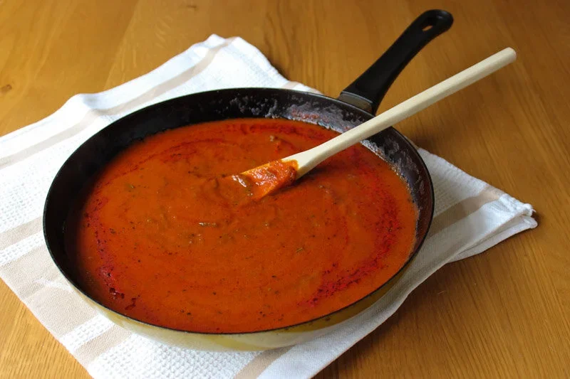 A frying pan filled with rich, smooth red tomato sugo sauce with a wooden spoon resting inside, set on a white tea towel.